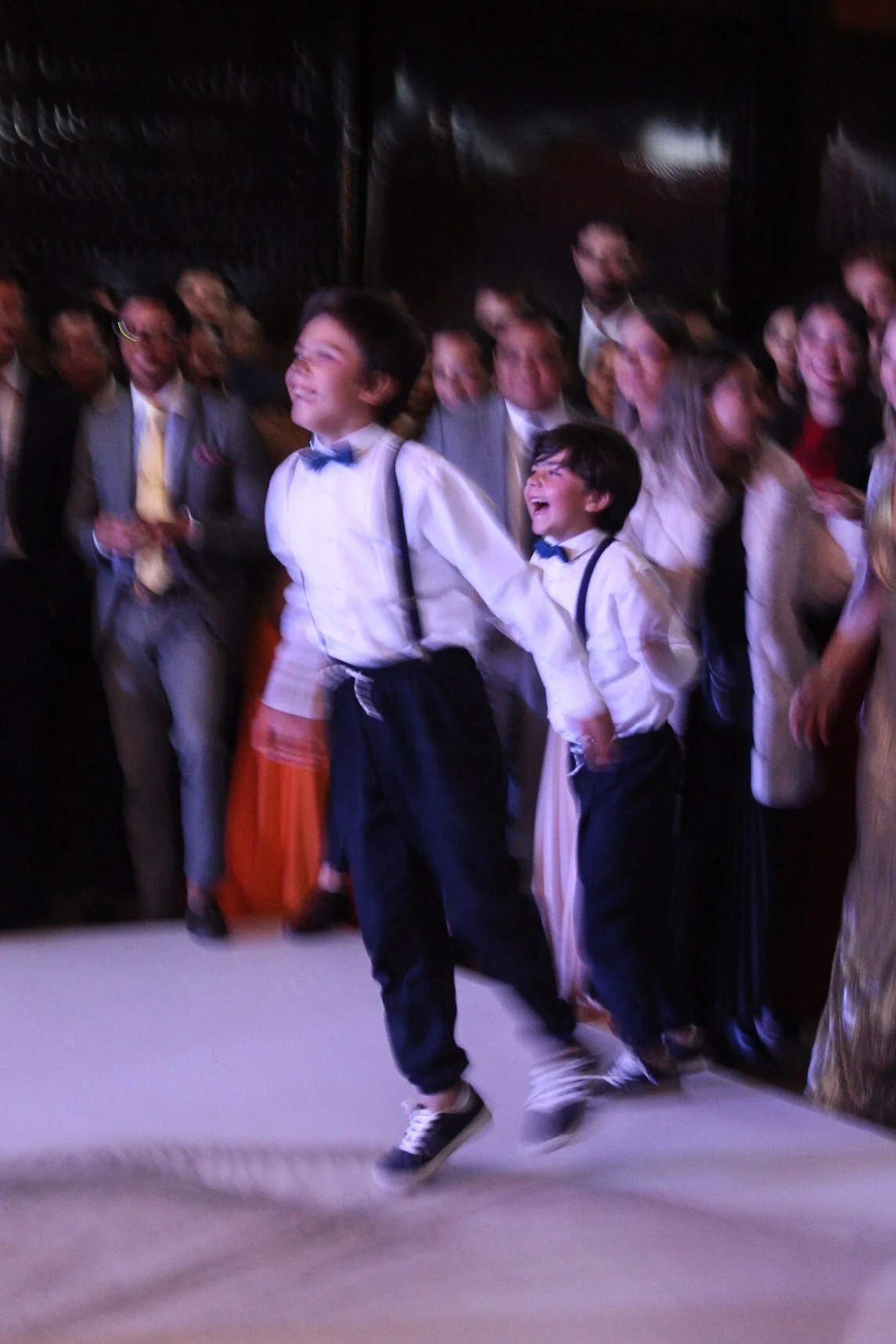 Dos niños niños en ropa formal y con corbata azul se están divirtiendo y bailando en una celebración con muchas personas en el fondo.