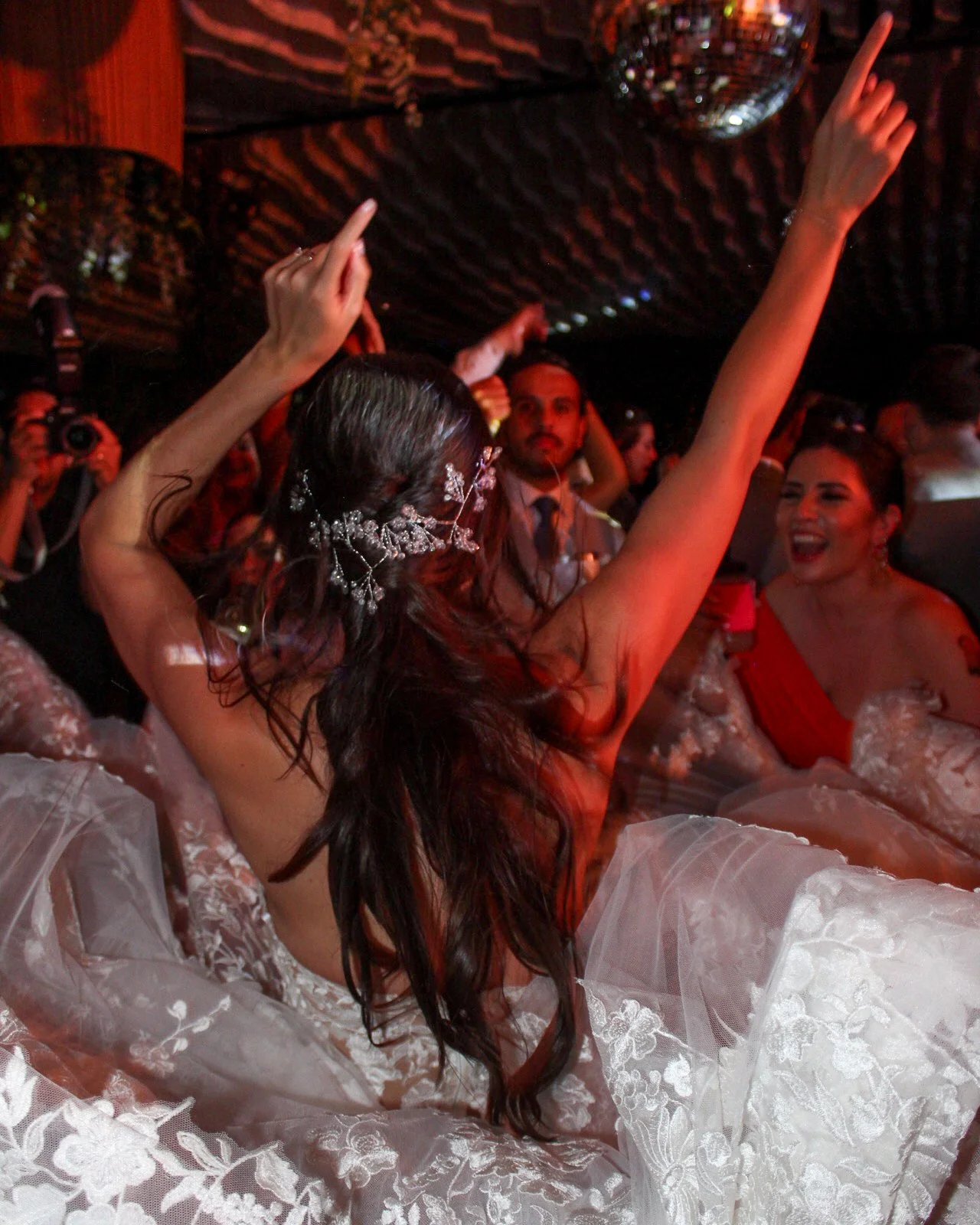 Una mujer en vestido de novia bailando en una fiesta, rodeada de otros invitados que también disfrutan del momento, en un ambiente festivo con luces y decoración.