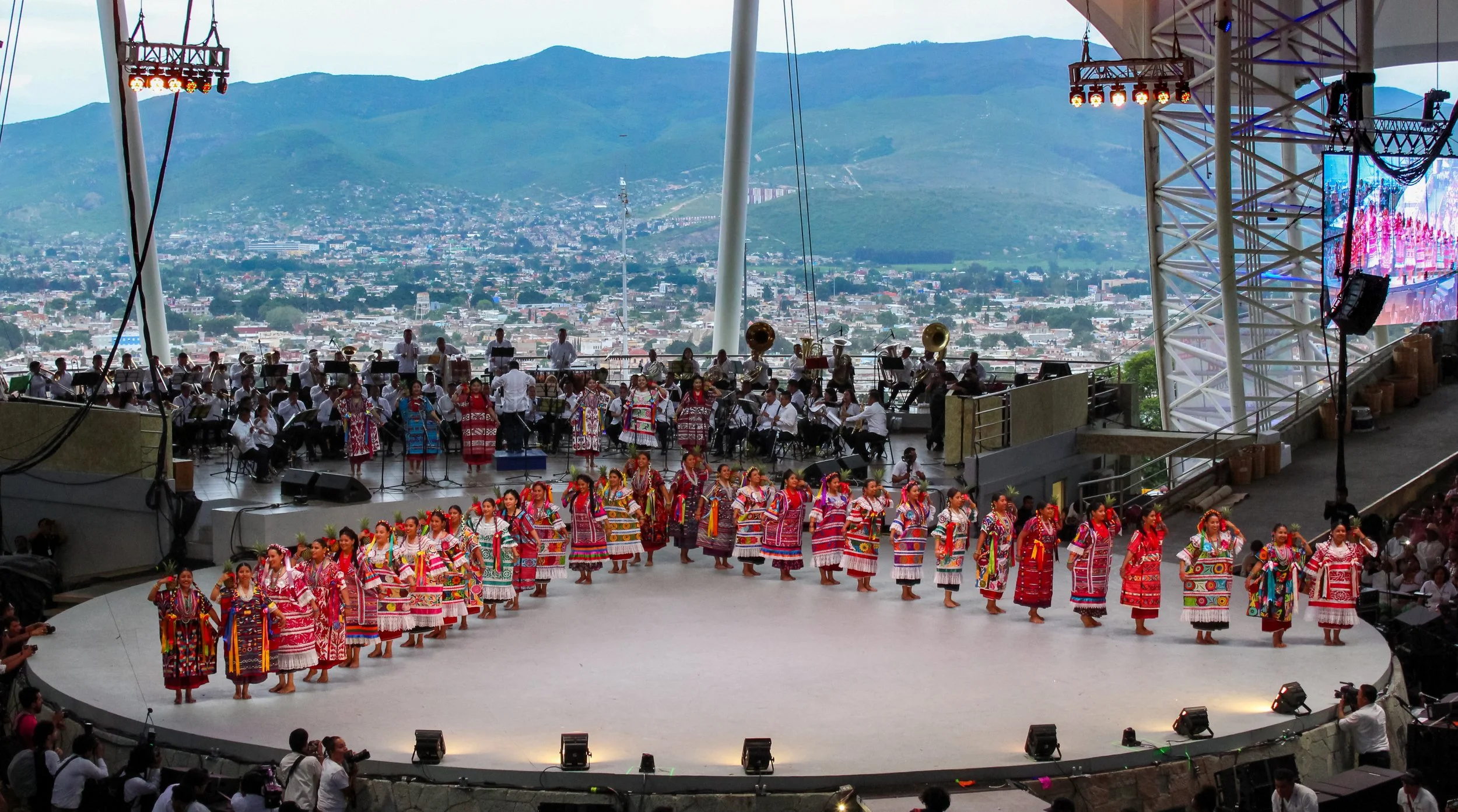 Una presentación de grupo de personas con trajes tradicionales en un escenario circular, con una orquesta y una ciudad de fondo en las montañas.