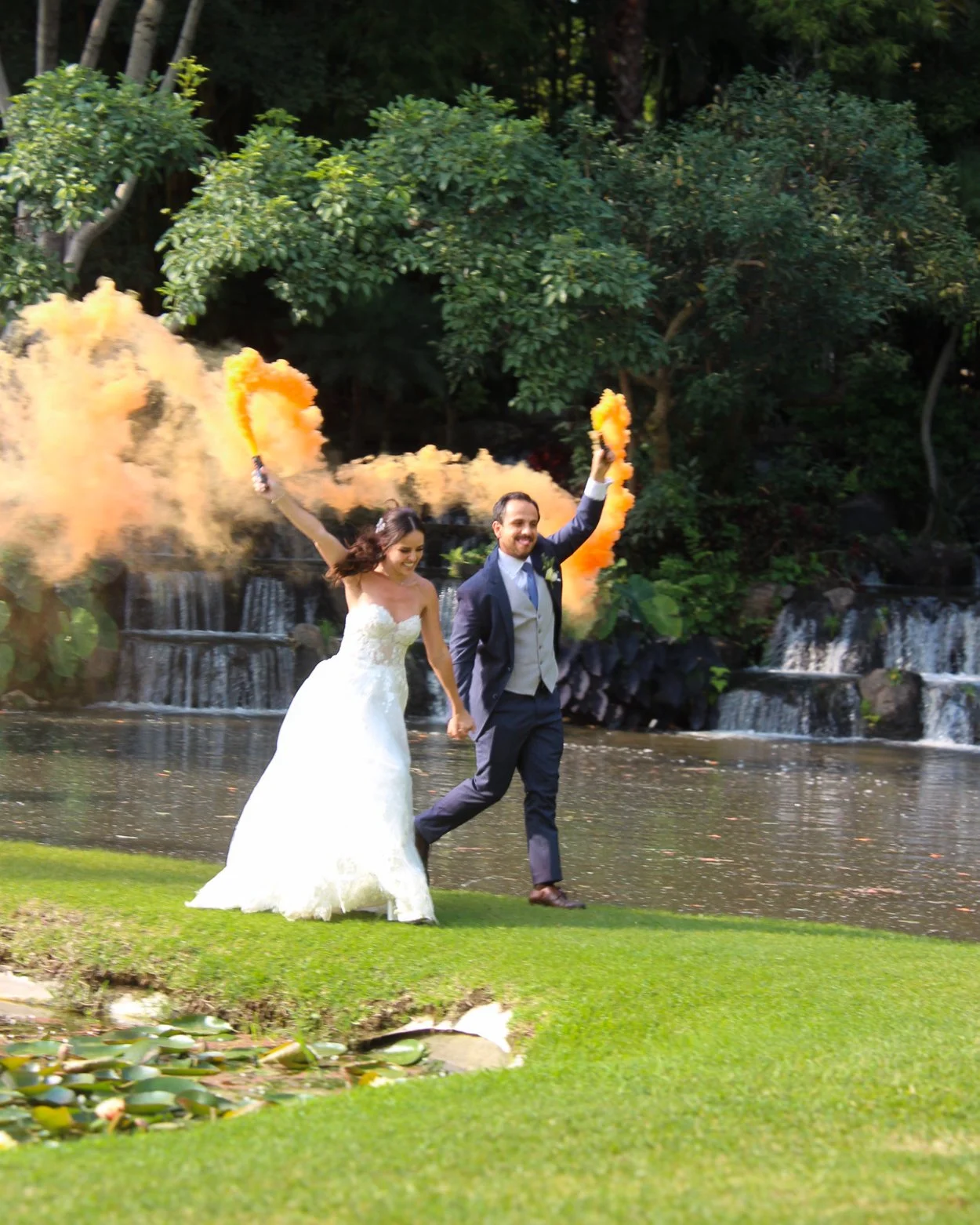 Pareja de boda, mujer en vestido blanco y hombre en traje oscuro, corriendo en un paisaje natural con agua y árboles, lanzando humo de color naranja.