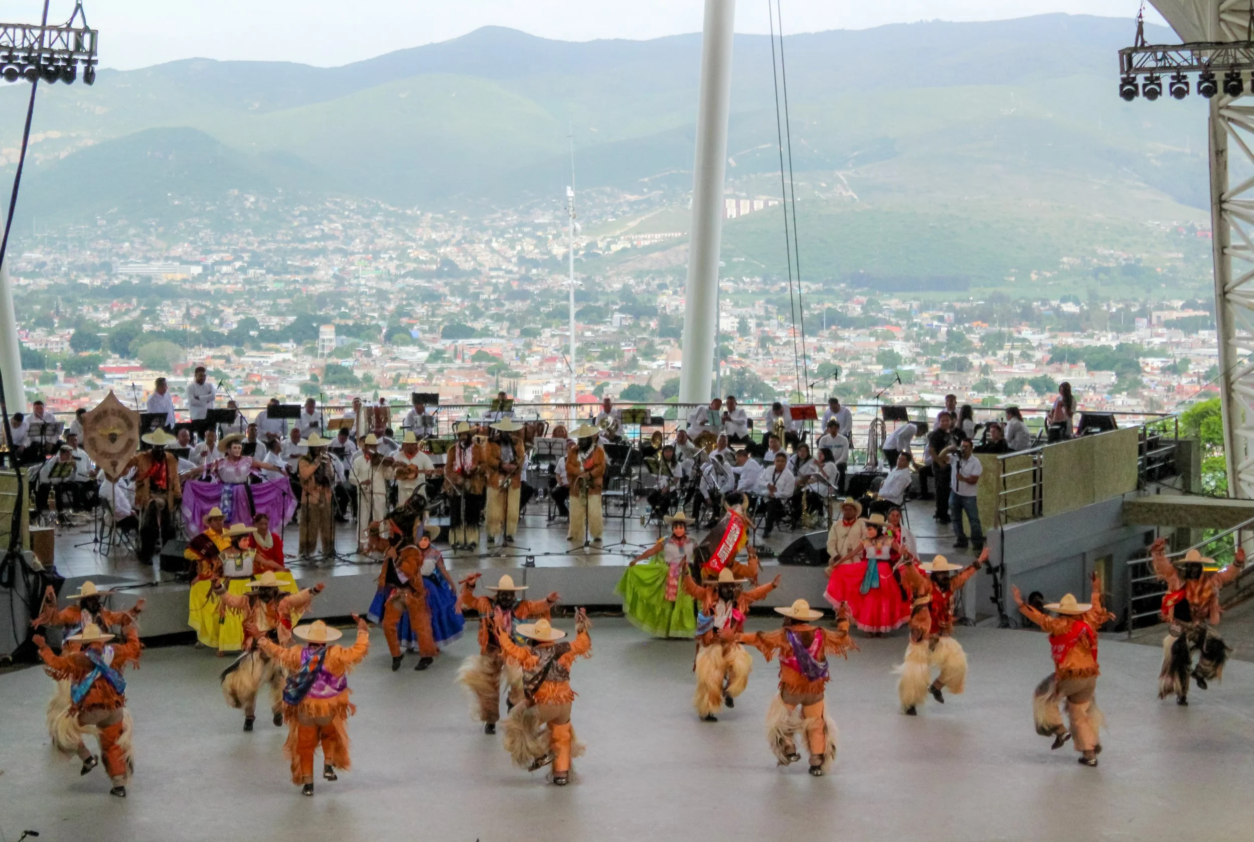 Danza folclórica con bailarines con trajes tradicionales en un escenario, acompañada por una orquesta en vivo, con una vista panorámica de una ciudad y montañas al fondo.