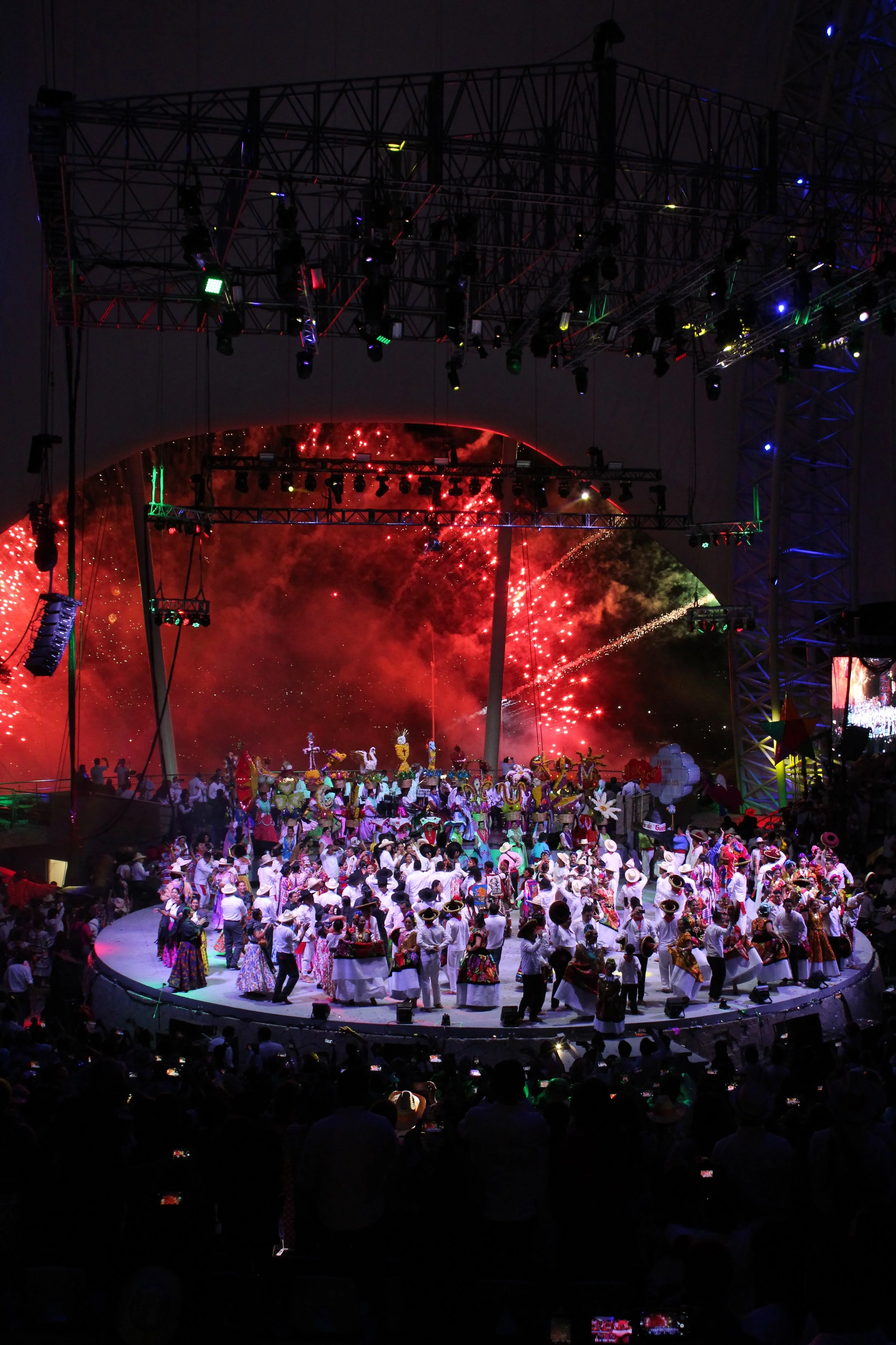 Escenario con una celebración tradicional, con muchas personas vestidas con ropa folklórica y fuegos artificiales en el fondo.