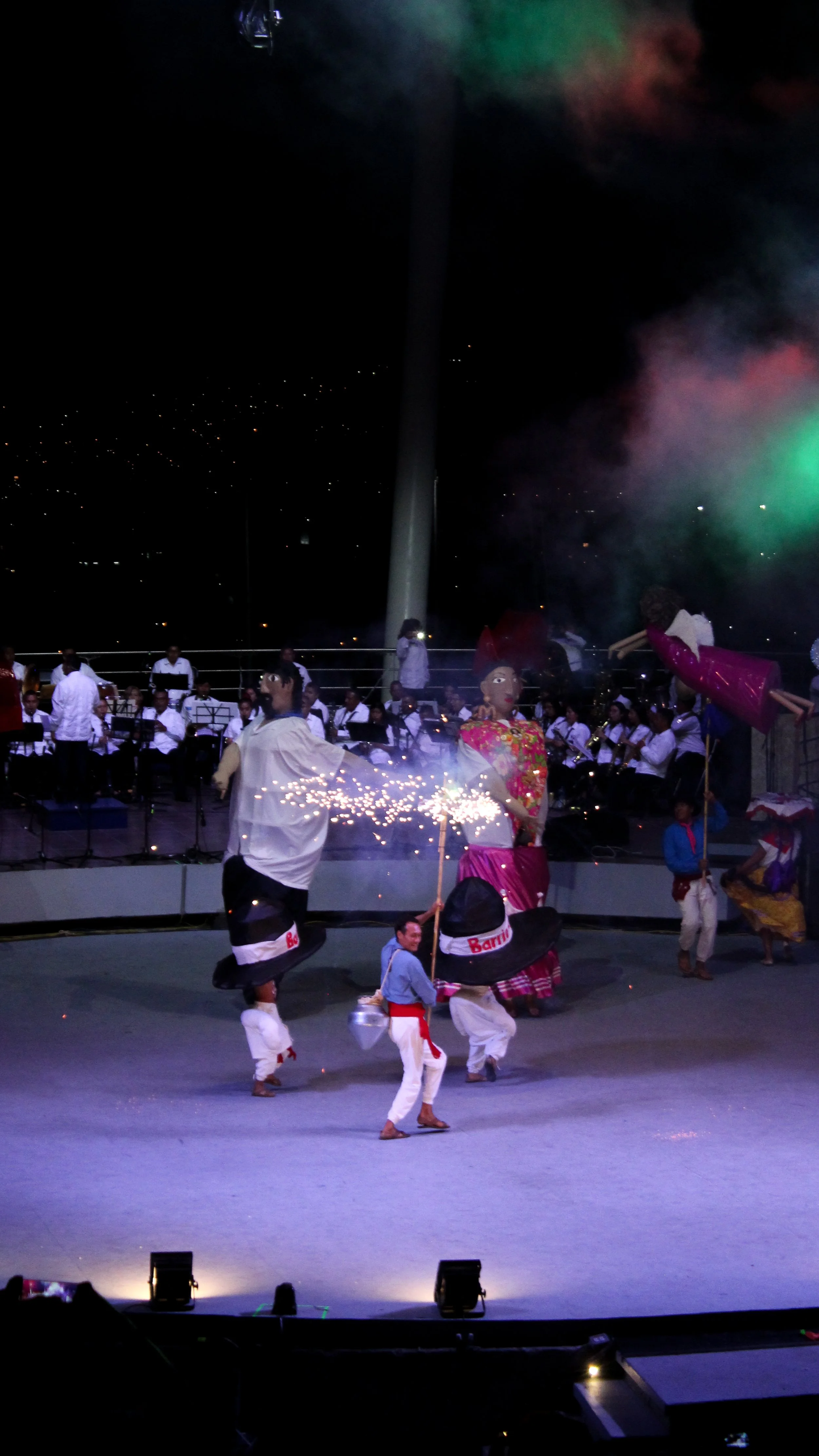 Escenario con bailarines tradicionales, música en vivo, y fuegos artificiales, en una celebración cultural nocturna.