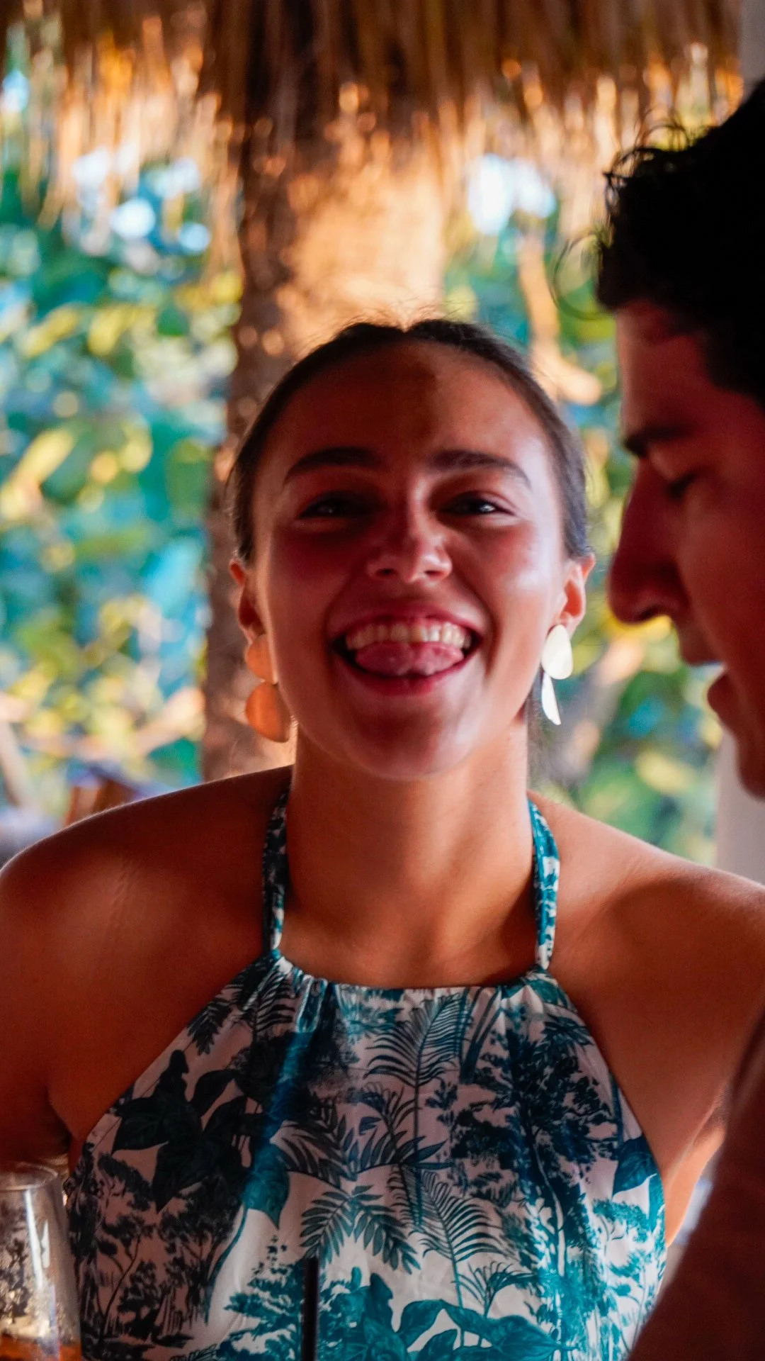 Una mujer joven sonriendo y haciendo una cara feliz, con un fondo de árboles y naturaleza, en un entorno tropical o de playa, con otra persona parcialmente visible a su lado.