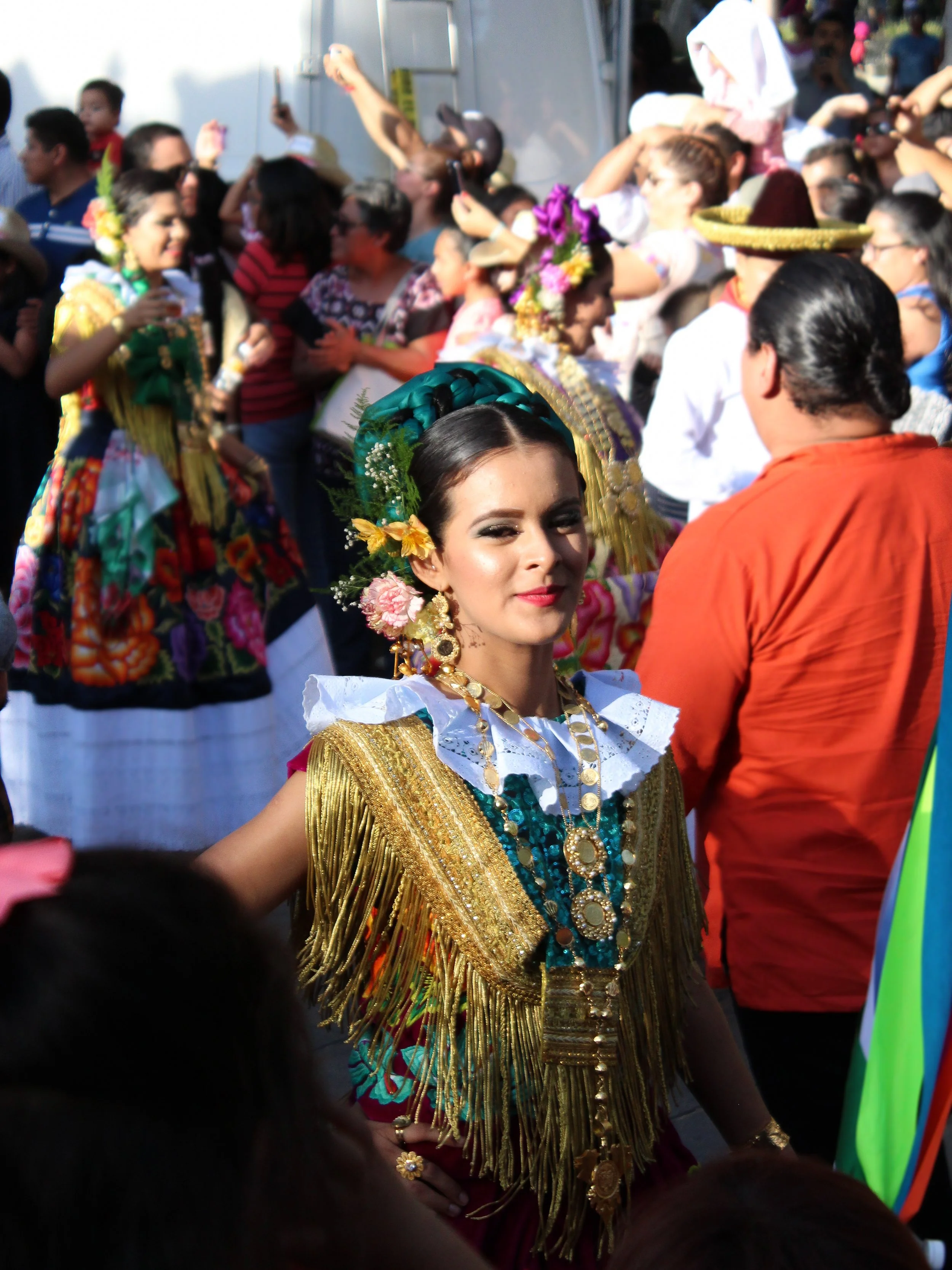 Mujer vestida con ropa tradicional mexicana y accesorios de oro en un evento cultural con muchas otras personas en el fondo.