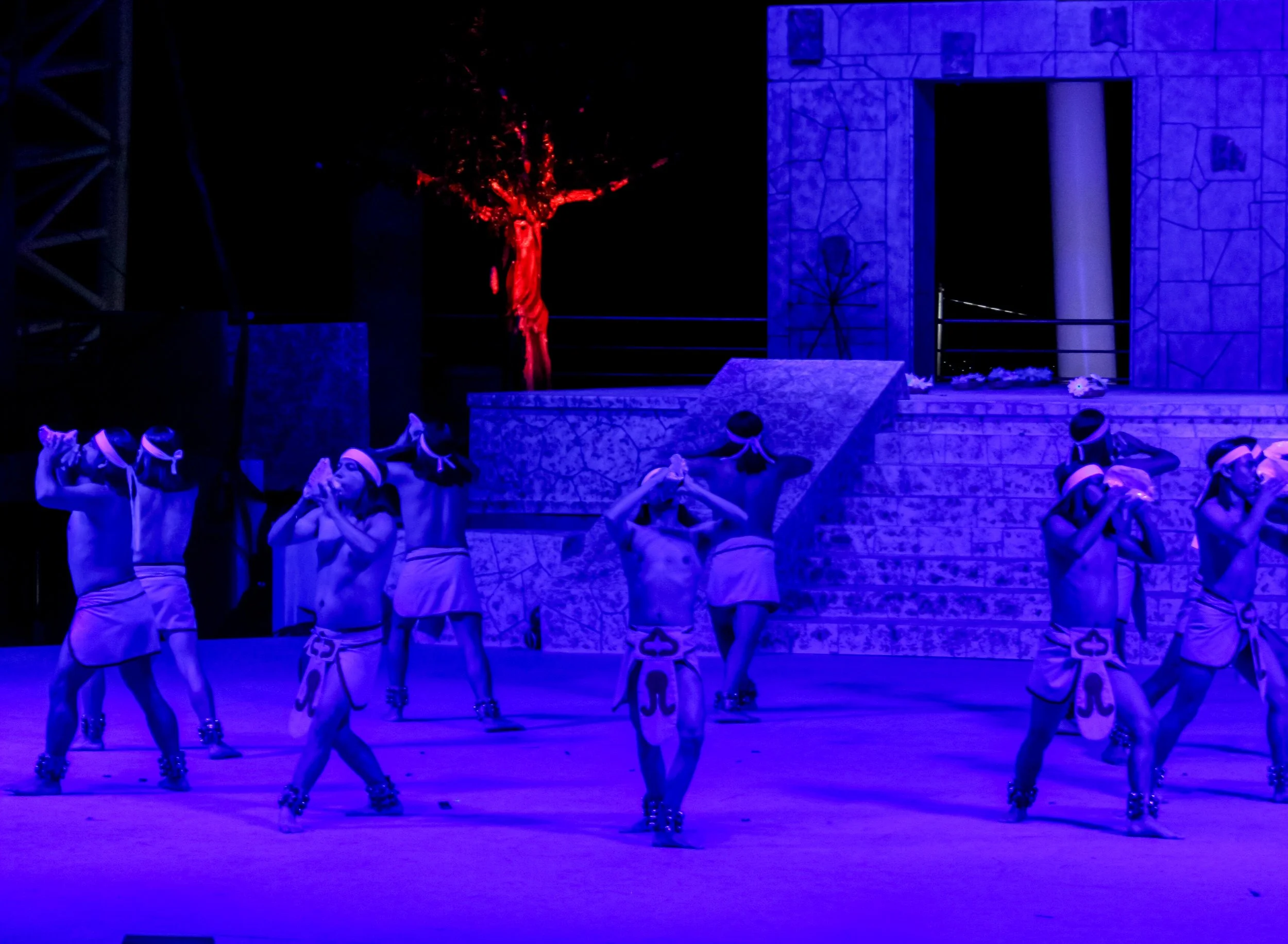 Bailarines con atuendo tradicional en una actuación bajo luz azul, en un escenario con escaleras y decoraciones de estilo antiguo, incluyendo una gran cruz y un árbol iluminado en rojo.