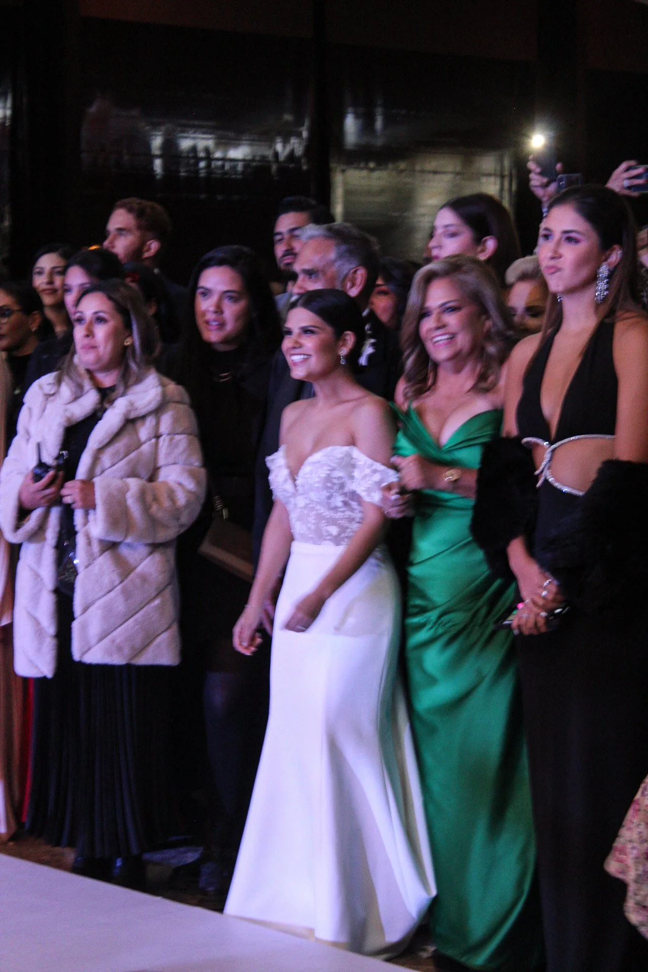 Grupo de personas en un evento formal, algunas sonriendo y vestidas con trajes elegantes, en un ambiente de celebración.