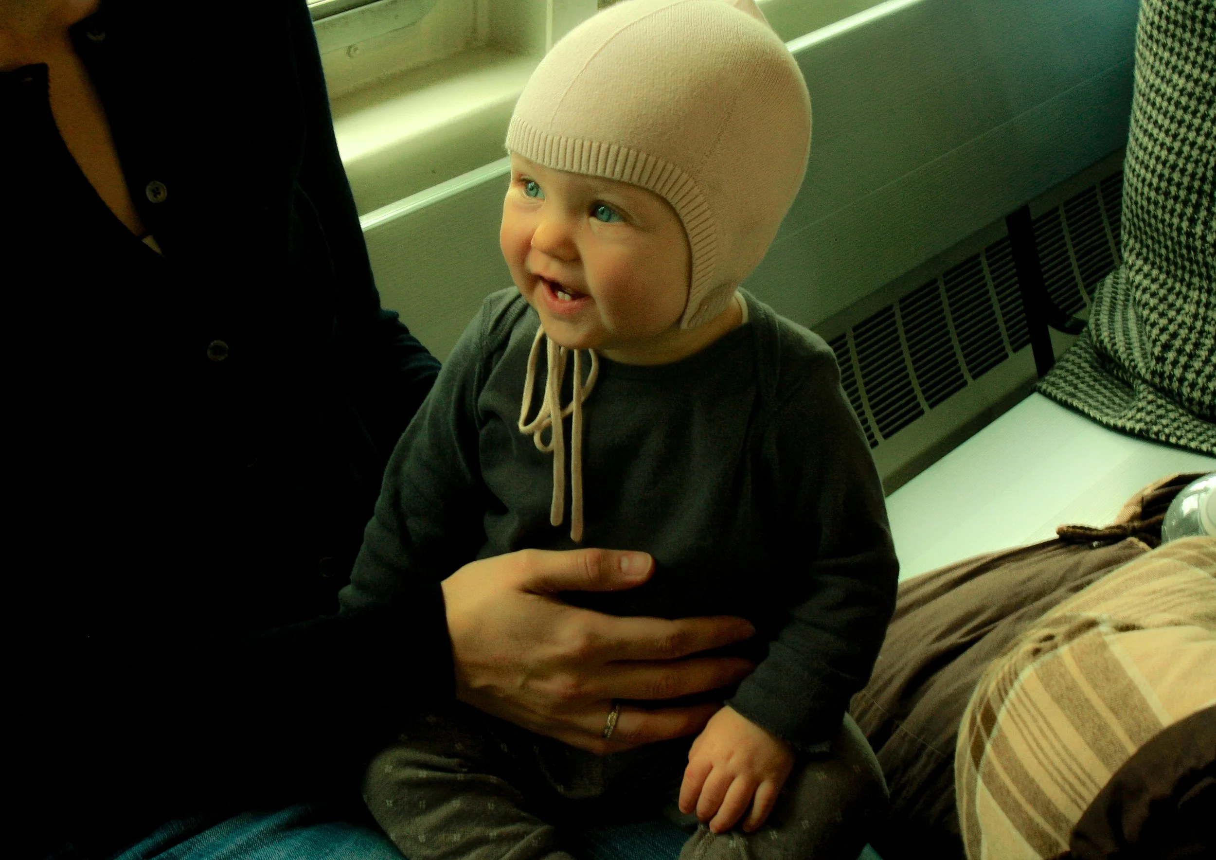 Niño pequeño con gorro de color beige sentado sobre las piernas de un adulto, sonriendo y mostrando dientes. Está en un ambiente interior, junto a una ventana y una unidad de calefacción.