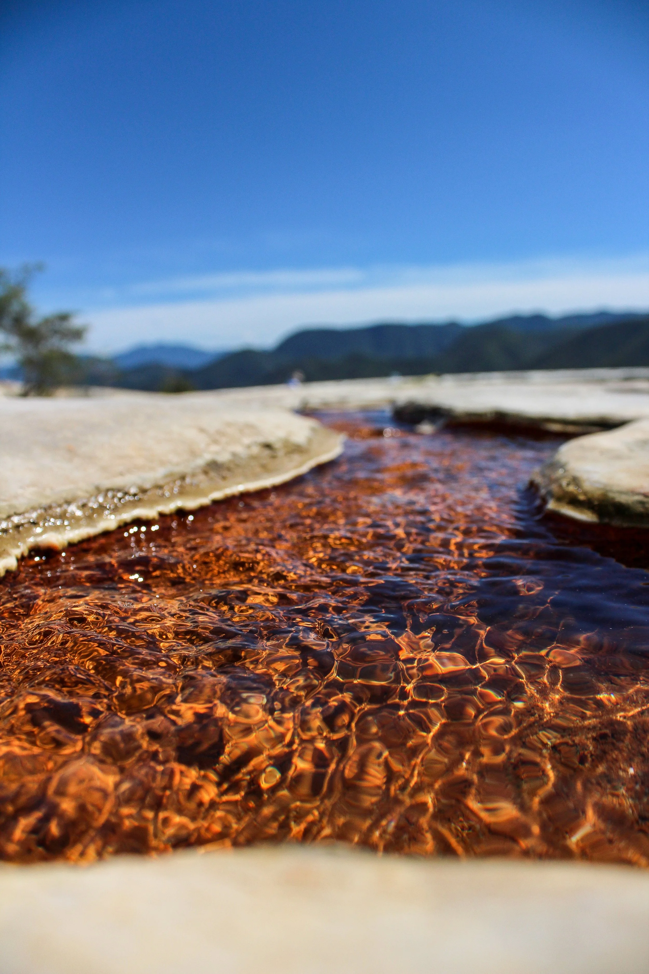 Un río de agua clara y rojiza en un paisaje montañoso con cielo azul y poca vegetación.