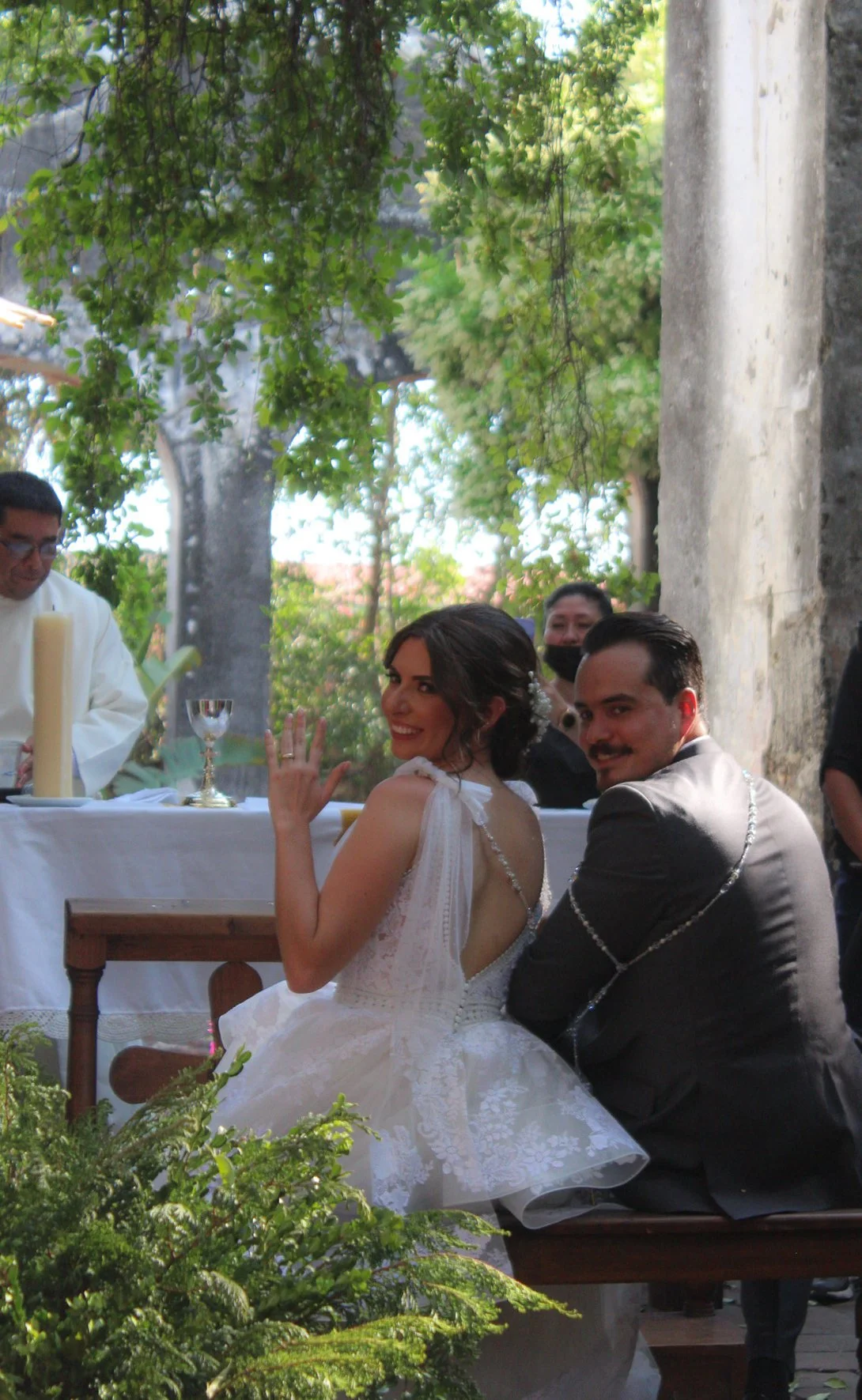Una pareja de novios en una boda, ella con vestido blanco y él con traje negro, sonrientes y sentados en un banco, rodeados de vegetación y con un altar al fondo donde un sacerdote realiza la ceremonia en un lugar al aire libre.