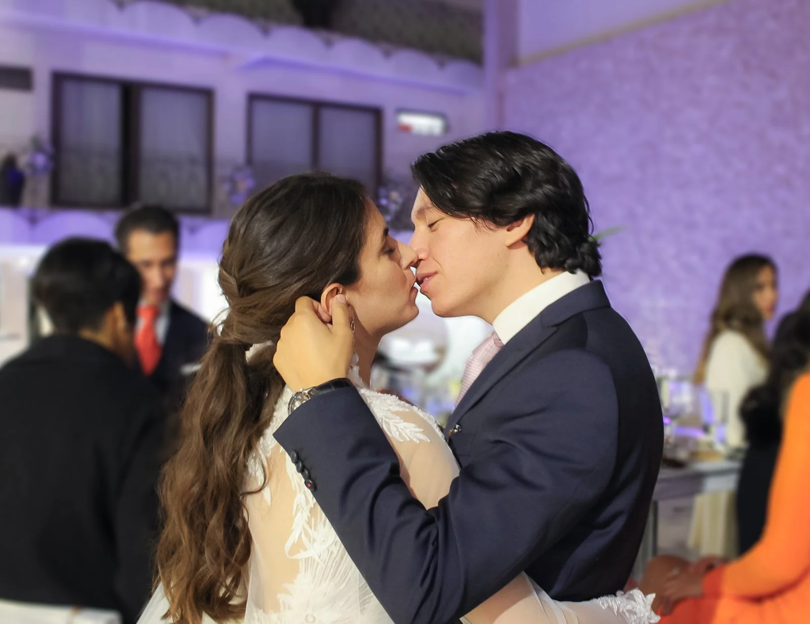 Pareja bailando y besándose en una boda, con fondo de invitados y decoraciones elegantes en un salón festivo.