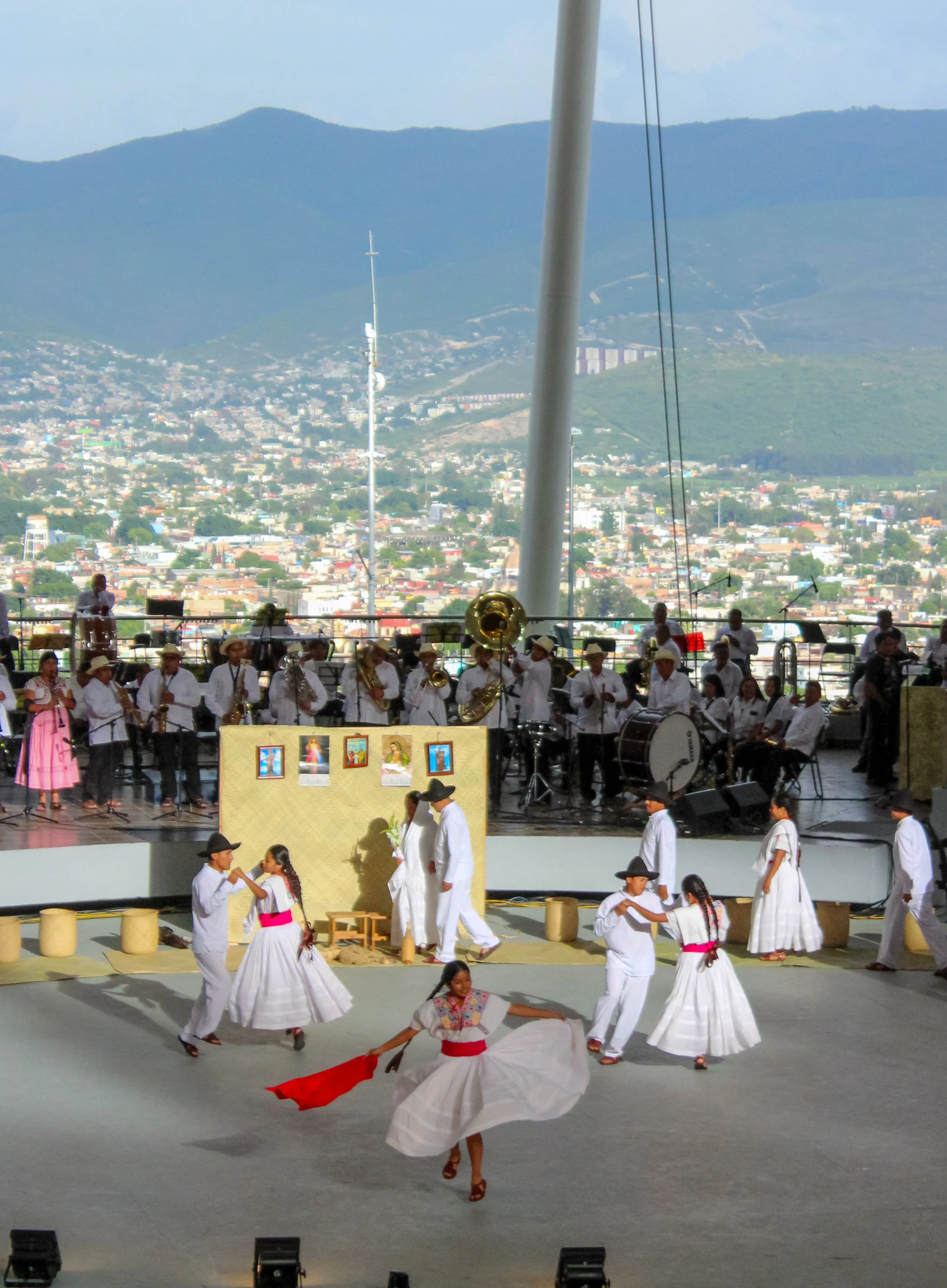 Un grupo de personas visten trajes tradicionales blancos y bailan en el escenario, mientras una orquesta toca música al fondo, con una vista panorámica de una ciudad y montañas en el horizonte.