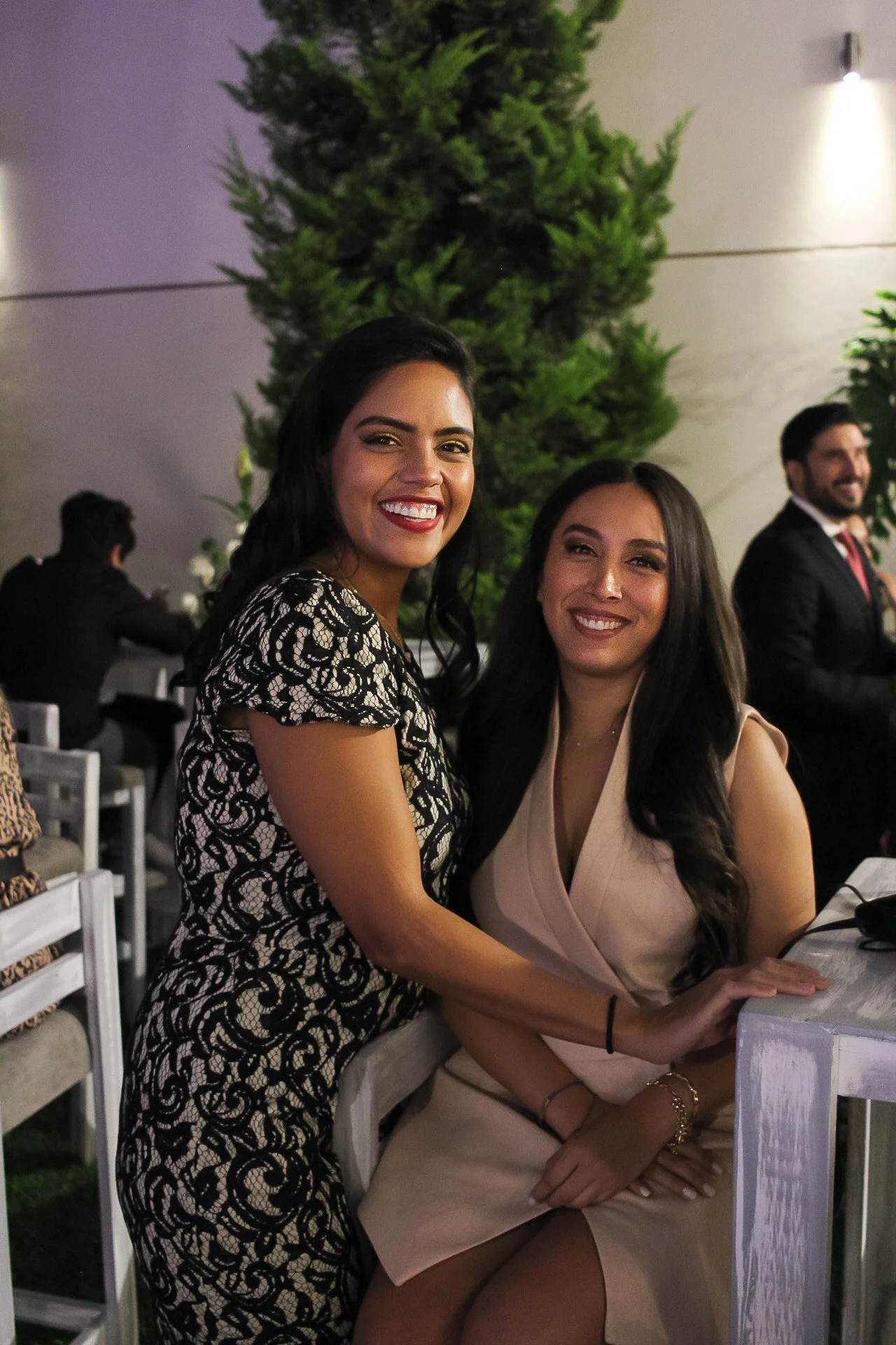 Dos mujeres sonriendo en un evento formal, una está de pie y la otra está sentada, con un fondo de plantas y otras personas en la celebración.
