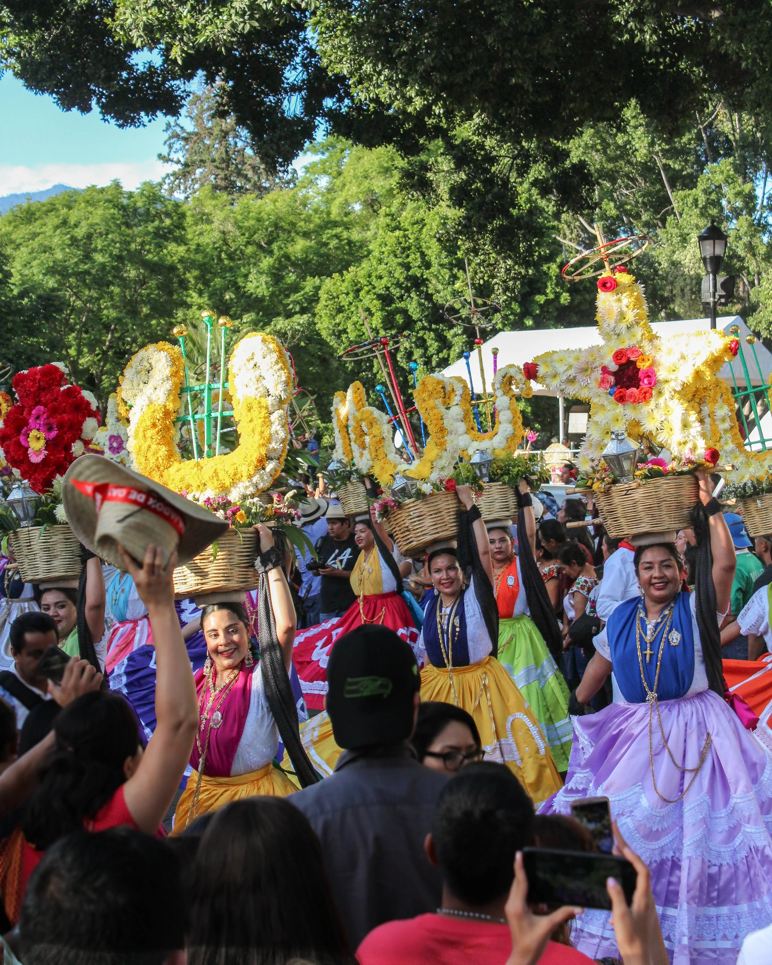 Personas vestidas con trajes tradicionales en un desfile, llevando cestas con decoraciones de flores en forma de figuras y objetos, en un parque con árboles verdes y cielo claro.
