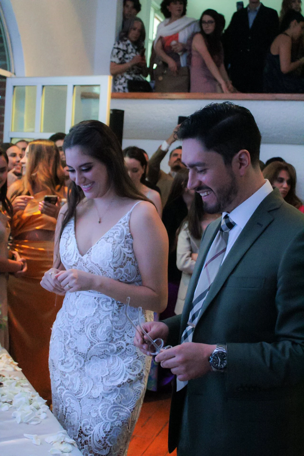 Pareja vestida formalmente en una celebración, la mujer lleva un vestido blanco de encaje y el hombre lleva traje y corbata, rodeados de varias personas en un lugar interior, probablemente una boda o evento especial.