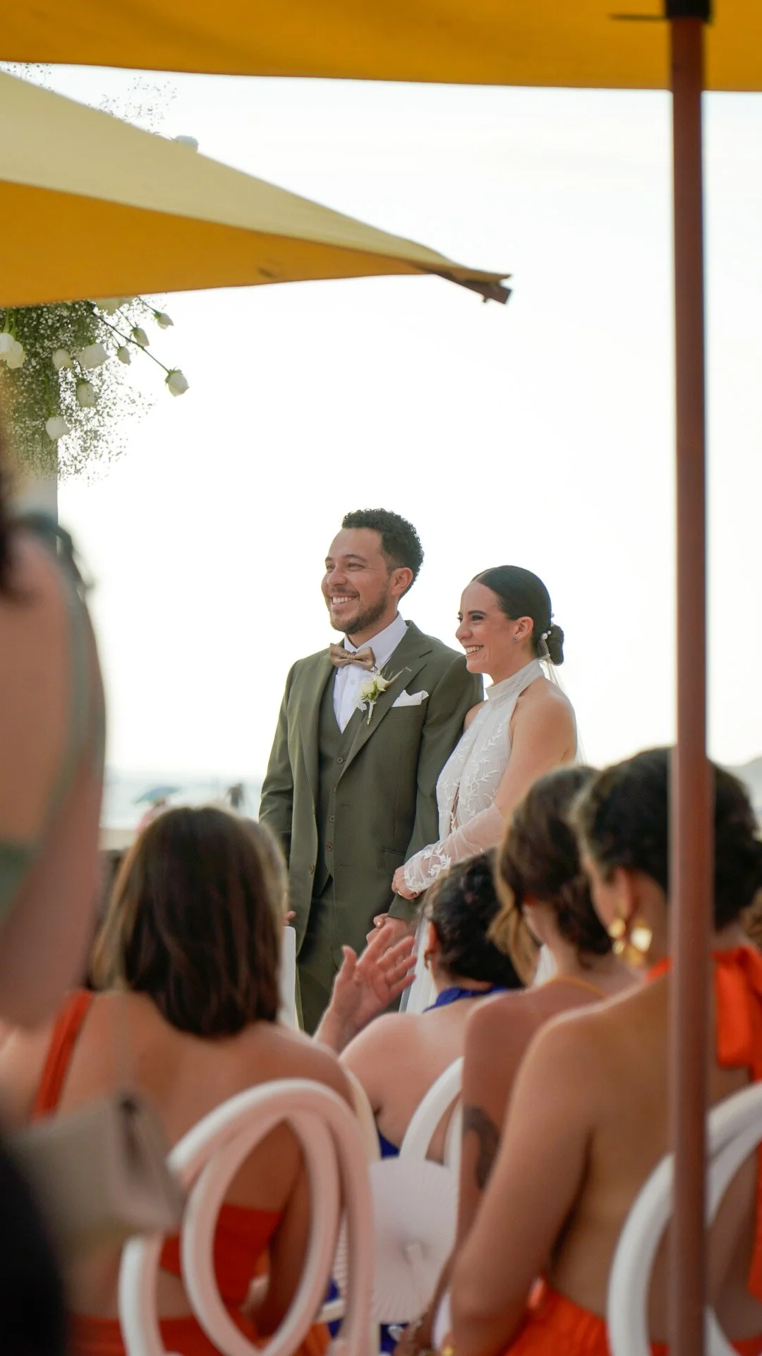 Pareja de recién casados en una ceremonia al aire libre, la mujer usa vestido blanco y el hombre traje gris con pajarita. Están sonriendo entre invitados con trajes y vestidos de colores vibrantes.