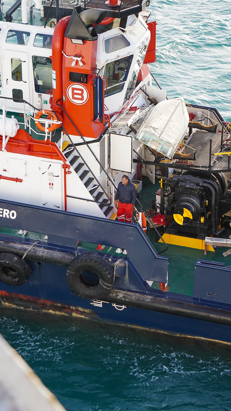 Un barco de rescate en el agua con dos personas en cubierta, una con pantalones rojos y chaqueta azul, y otra con pantalones rojos y chaqueta negra.
