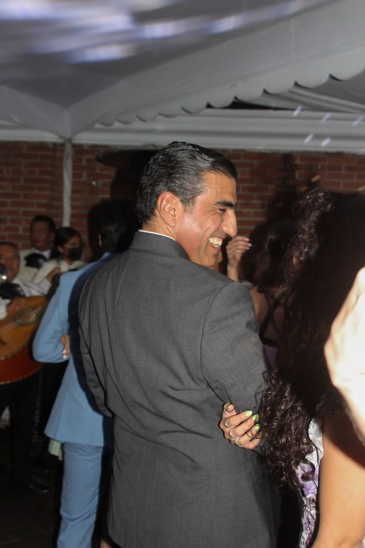 Hombre sonriendo en una reunión social, rodeado de otras personas, con paredes de ladrillo de fondo.