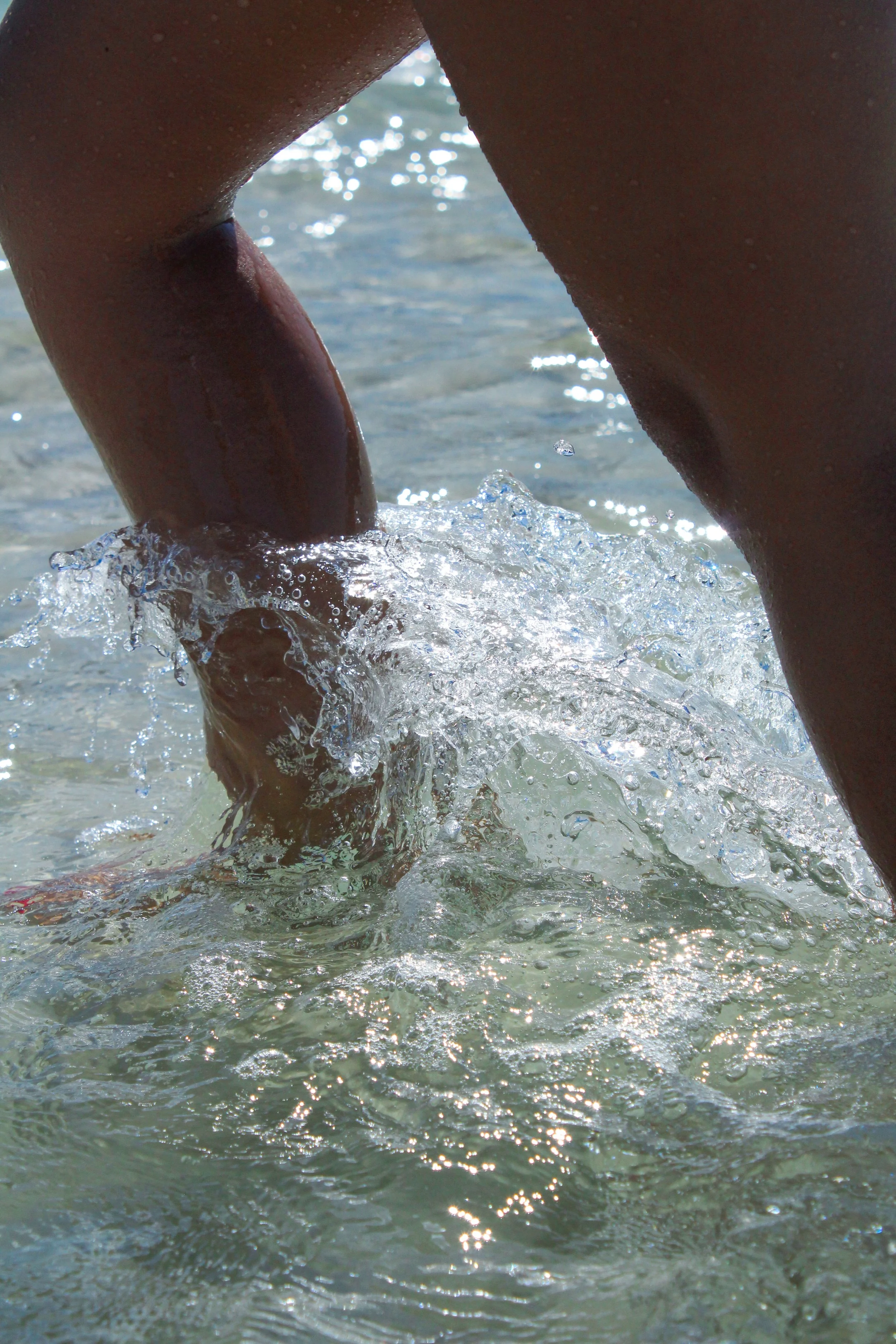 Imagen de unas piernas en el agua de la playa, con agua salpicando y reflejos de sol.