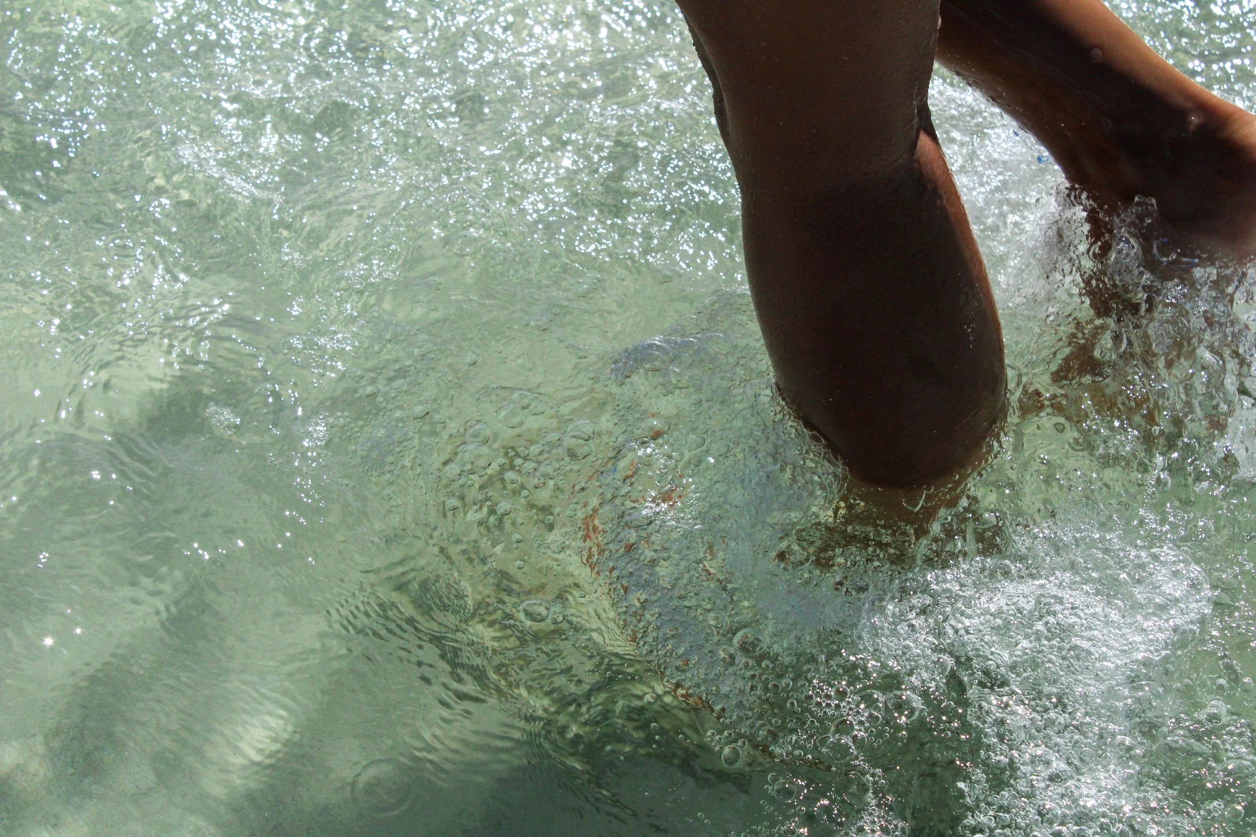 Persona en el agua, probablemente en la playa, realizando surf o natación, con enfoque en sus piernas y el agua a su alrededor.