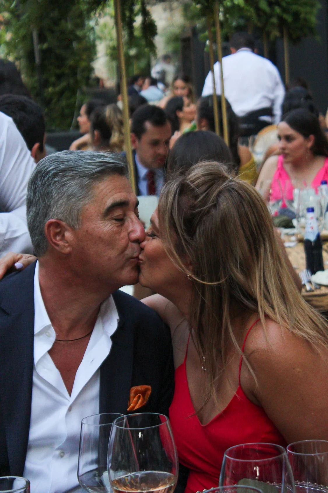 Una pareja se besa en una celebración al aire libre, rodeada de muchas personas sentadas en mesas con comida y bebidas.