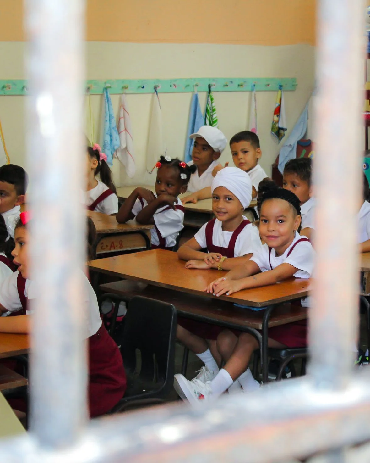 Niños en un aula escolar observando desde una reja, sentados en pupitres, algunos con uniformes y accesorios culturales.
