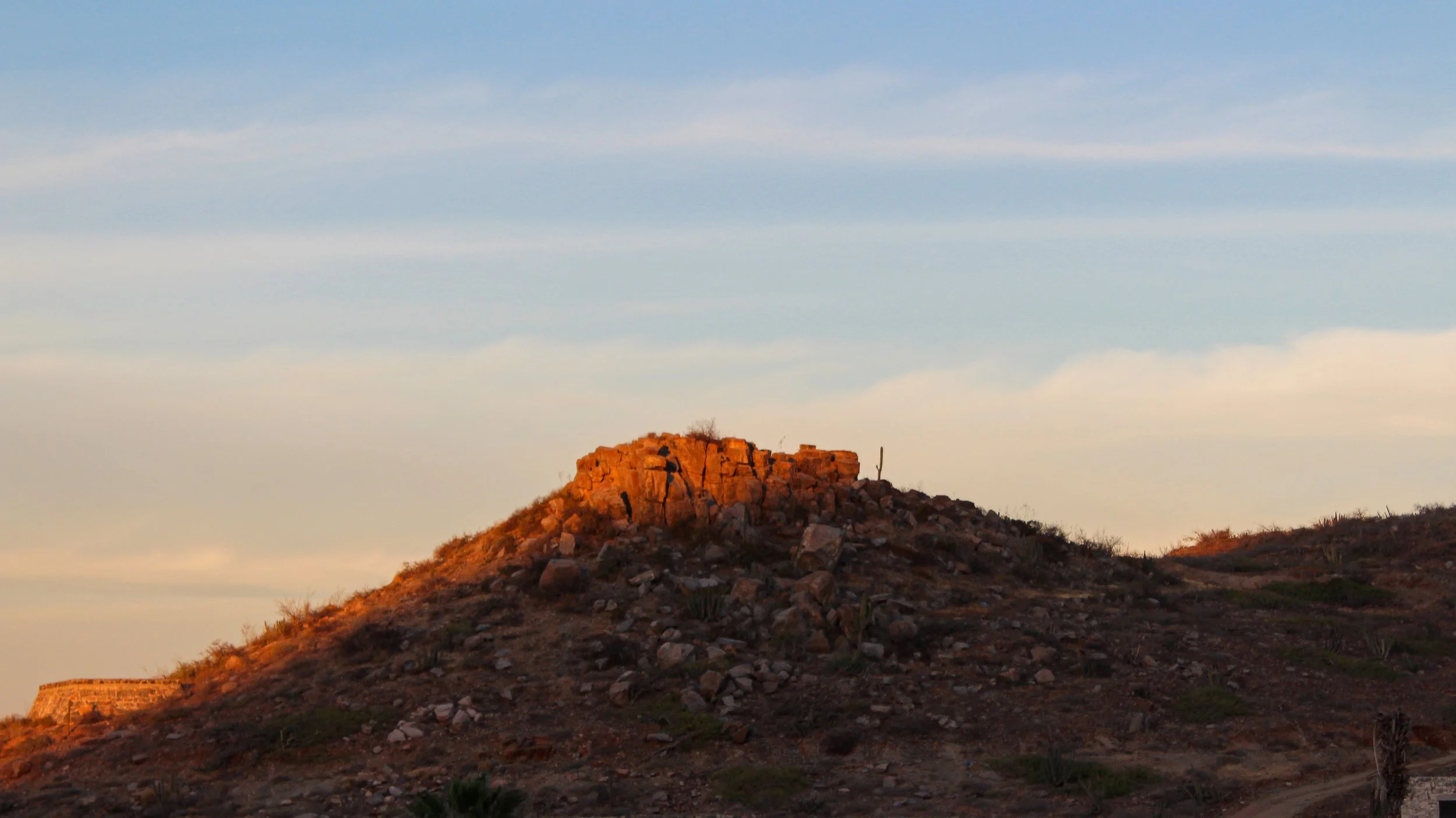 Montículo rocoso con ruinas antiguas y un árbol pequeño en la cima, en un paisaje árido con vegetación dispersa, bajo un cielo azul con nubes.