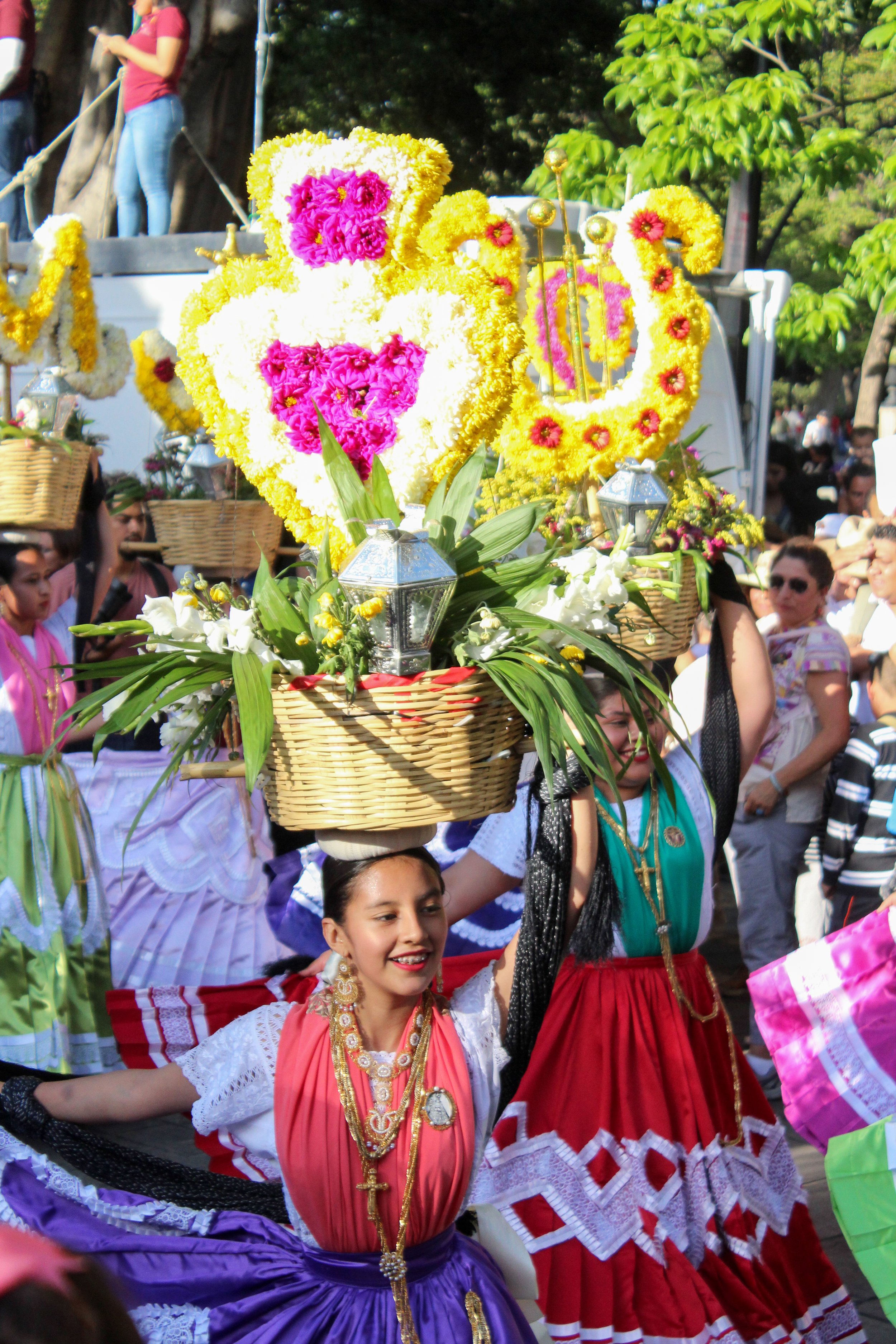 Mujeres vestidas con trajes tradicionales participando en un desfile, llevando canastas con decoraciones florales y cruces religiosas en la cabeza, rodeadas de un público observando.