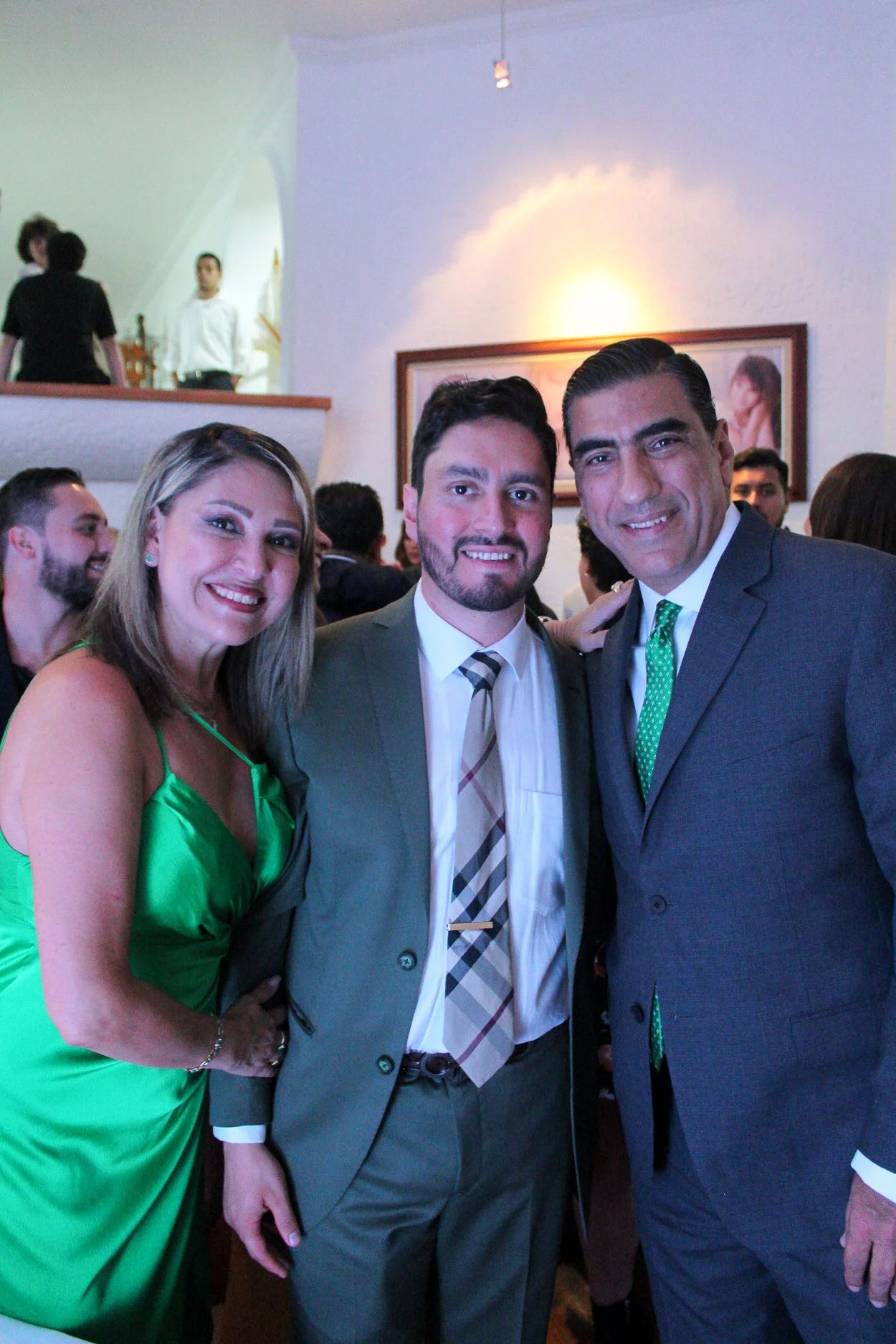 Tres personas en un evento social, son dos hombres y una mujer. La mujer lleva un vestido verde y los hombres llevan traje y corbata. Están sonriendo y posando para la foto.