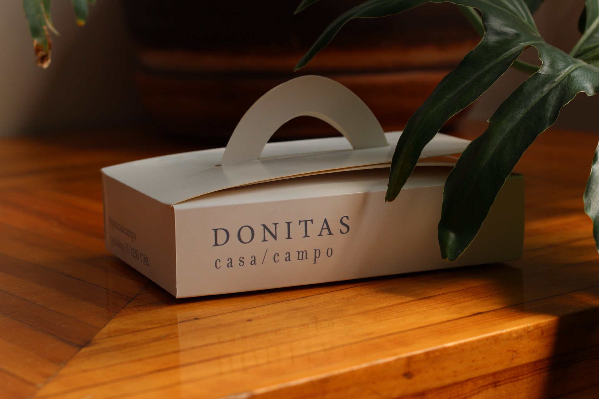 Caja de donitas con etiquetas 'casa/campo', en una mesa de madera, rodeada de hojas verdes.
