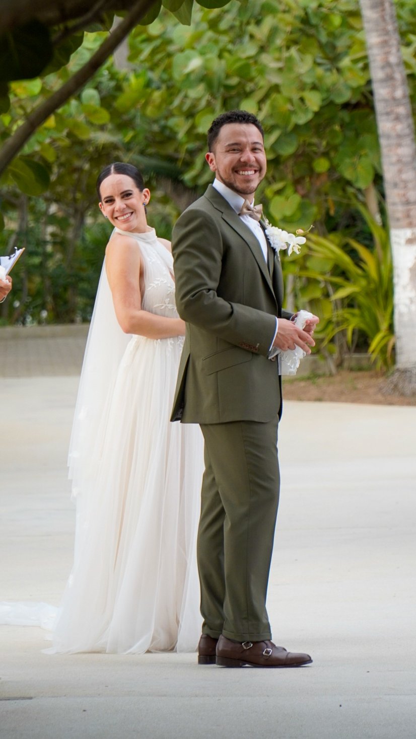 Pareja de novios sonriendo en una ceremonia en un entorno natural con árboles verdes y palmeras.