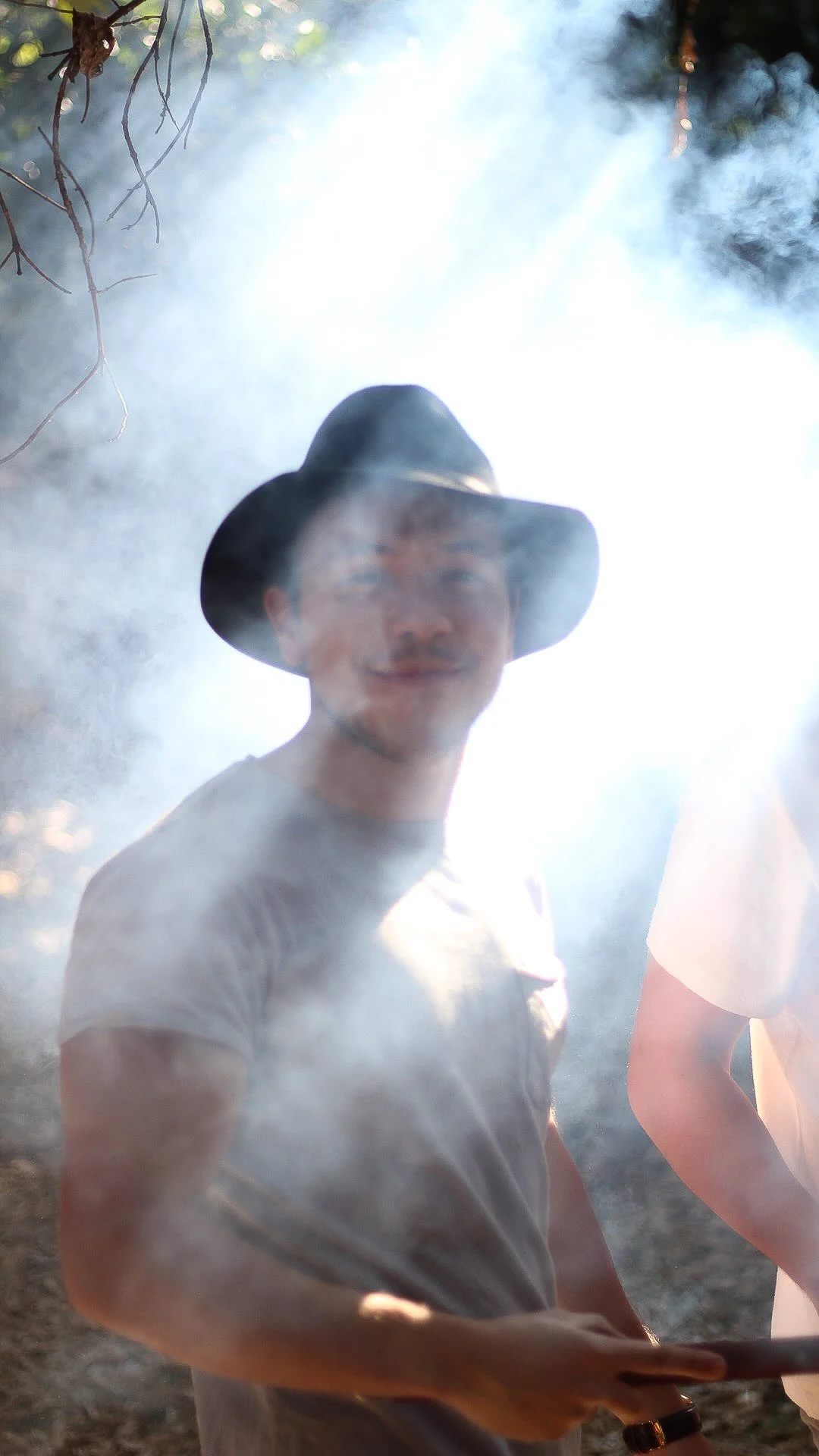 Joven con sombrero negro sonriendo en un entorno con humo y vegetación.