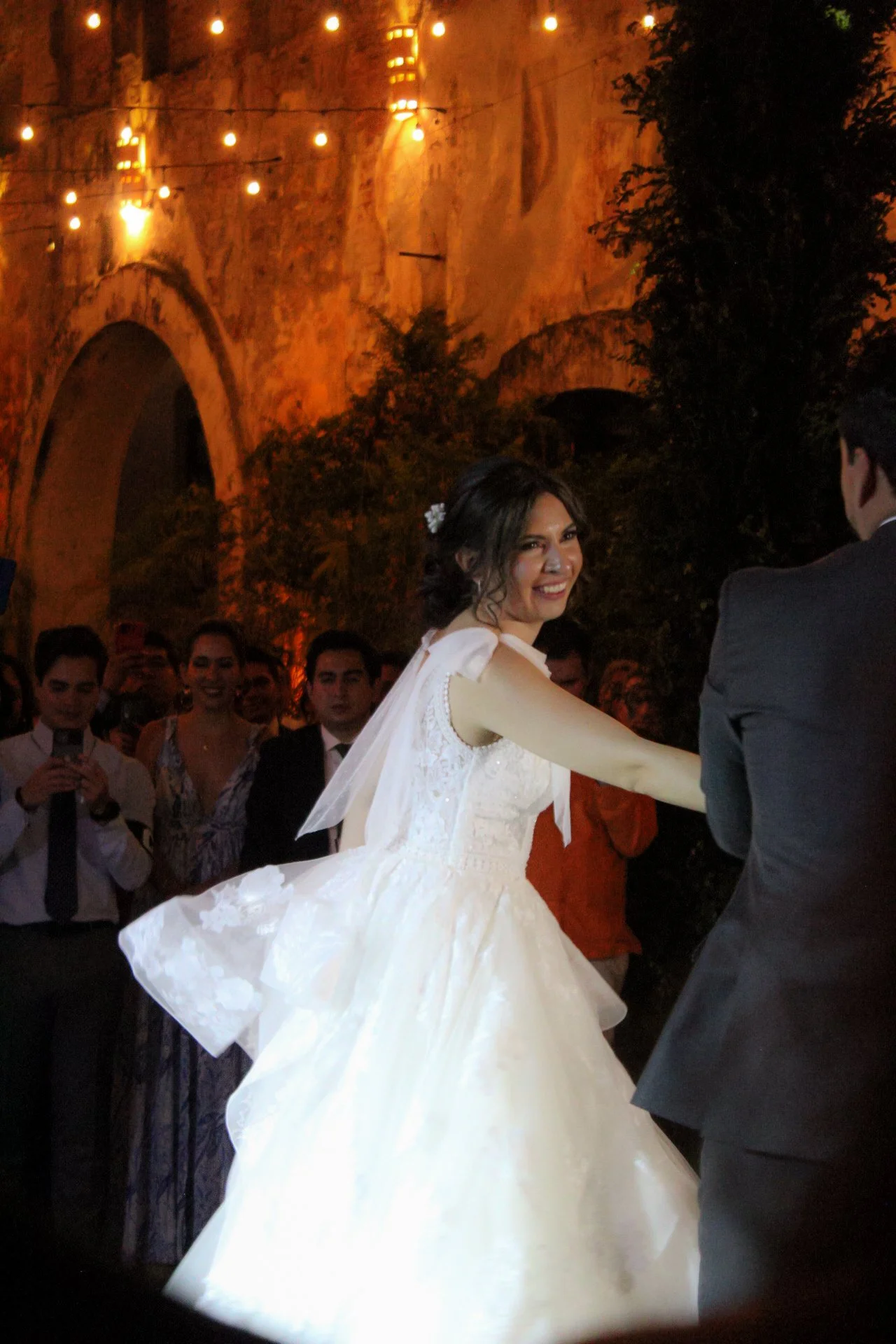 Una pareja bailando en una boda al atardecer, la novia vestida con un vestido de novia blanco y el novio con un traje oscuro, rodeados de invitados y decoración con luces.