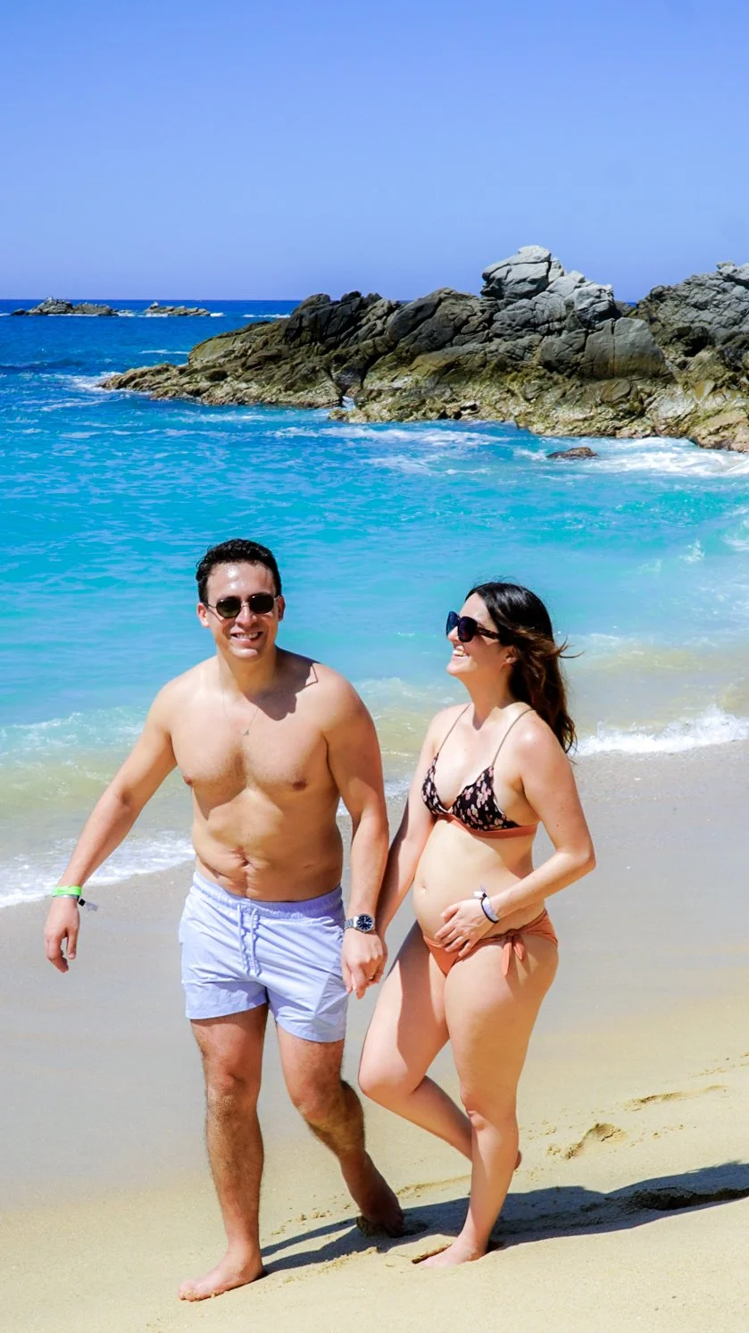 Una pareja caminando en la playa, con agua azul y rocas en el fondo, bajo un cielo despejado.