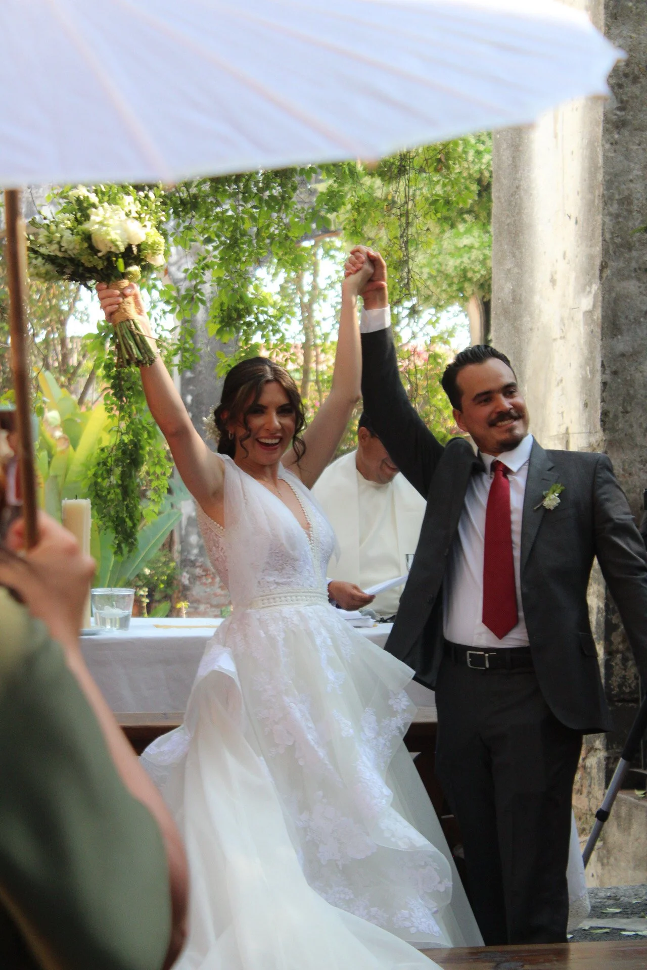Pareja de recién casados en una boda, levantando sus manos entrelazadas y sonriendo, en un entorno al aire libre con árboles y una pared de piedra, con invitados en el fondo y un oficiante.