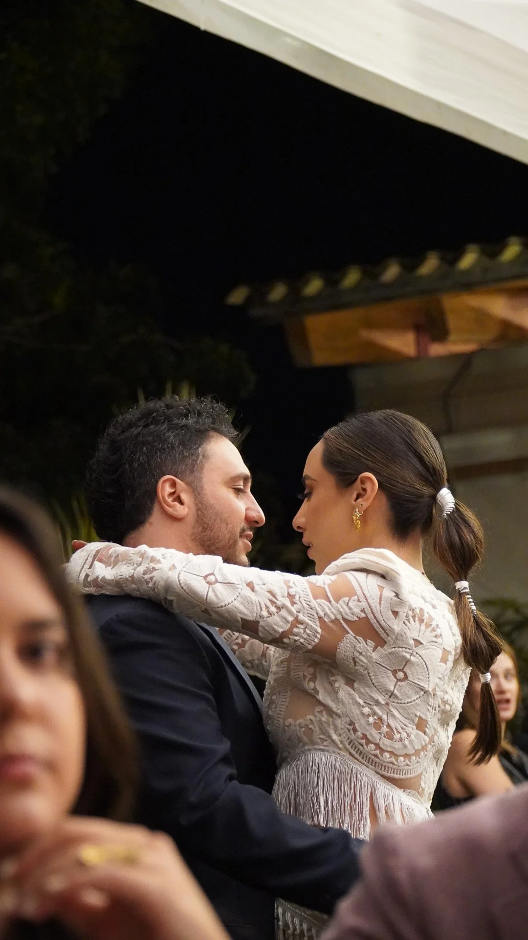 Pareja en un baile, ella con un vestido de encaje y pantalones de cabello castaño oscuro atado en una coleta, él con un saco oscuro y camisa blanca, compartiendo un momento romántico en una celebración.