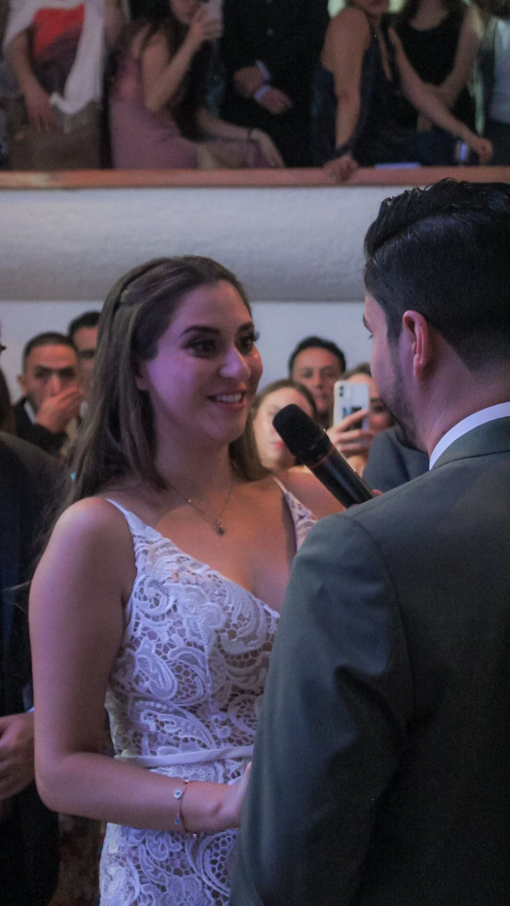 Una pareja en una ceremonia, la mujer sonríe mientras un hombre con micrófono la mira, en un entorno con otras personas.
