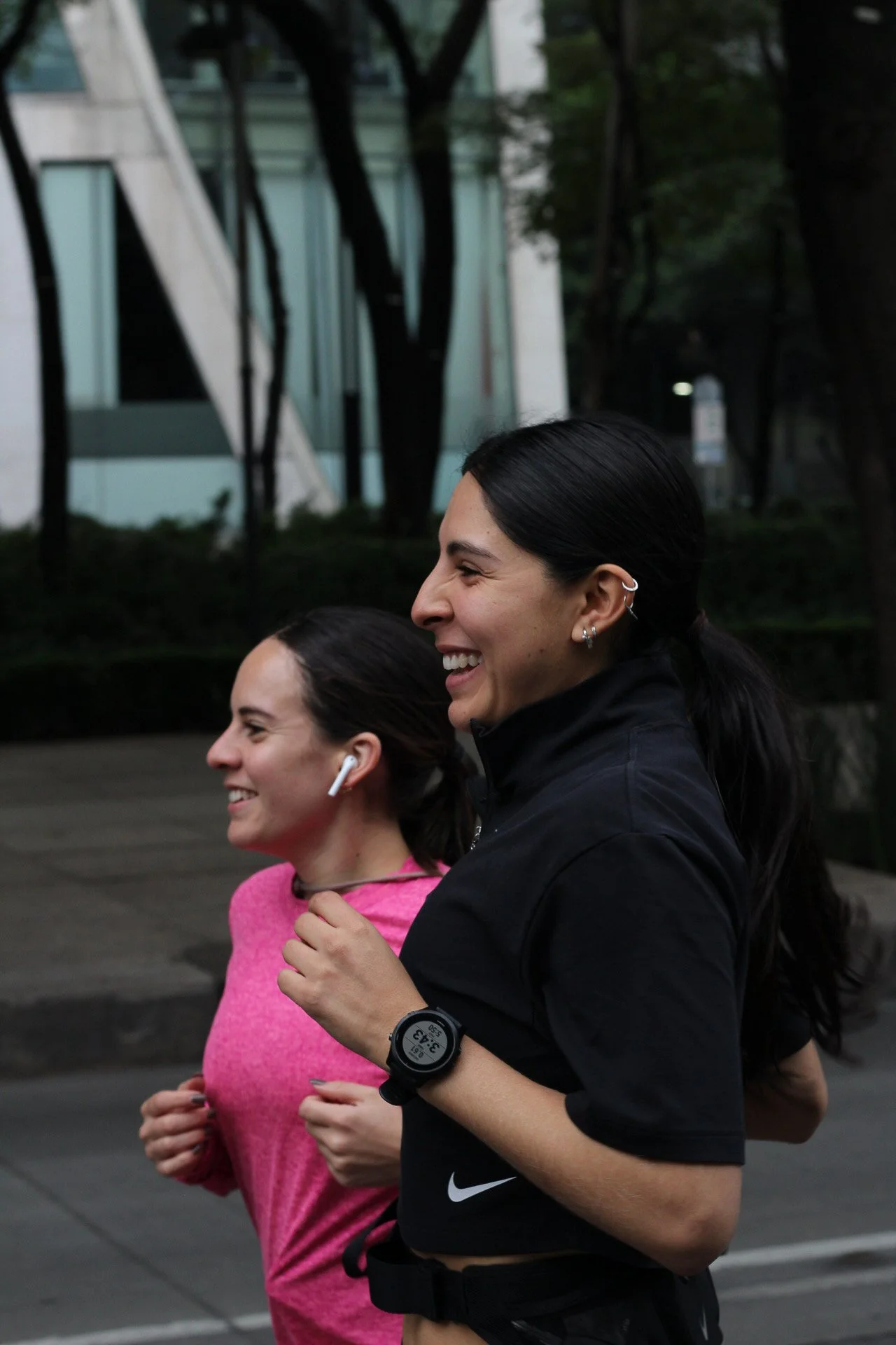 Dos mujeres corriendo en la calle, sonriendo y usando ropa deportiva, con fondo de árboles y edificios.