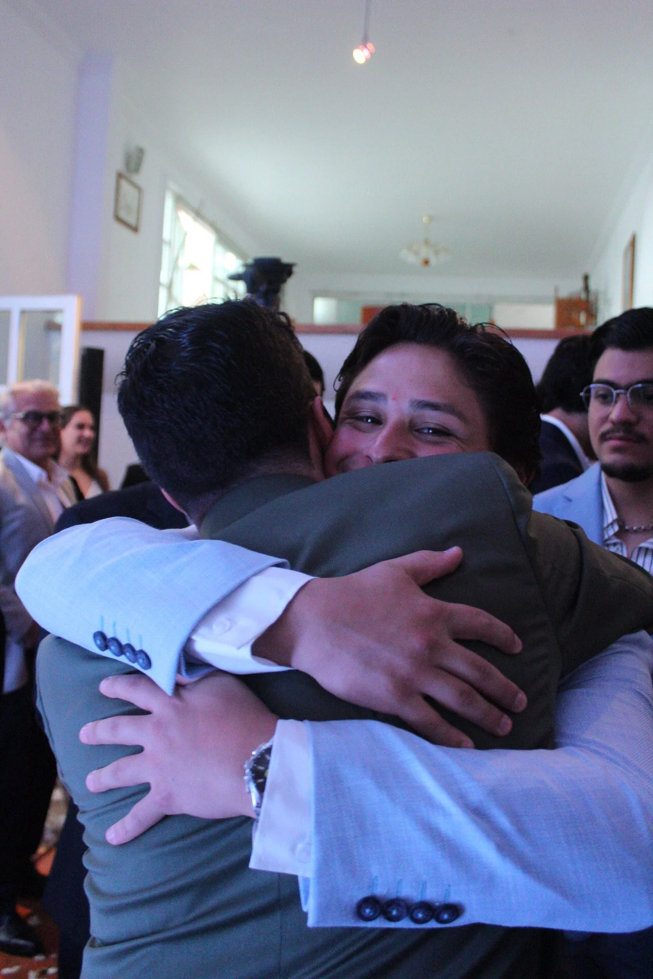 Dos hombres abrazándose, uno con sonrisa en un evento social con varias personas más en el fondo.