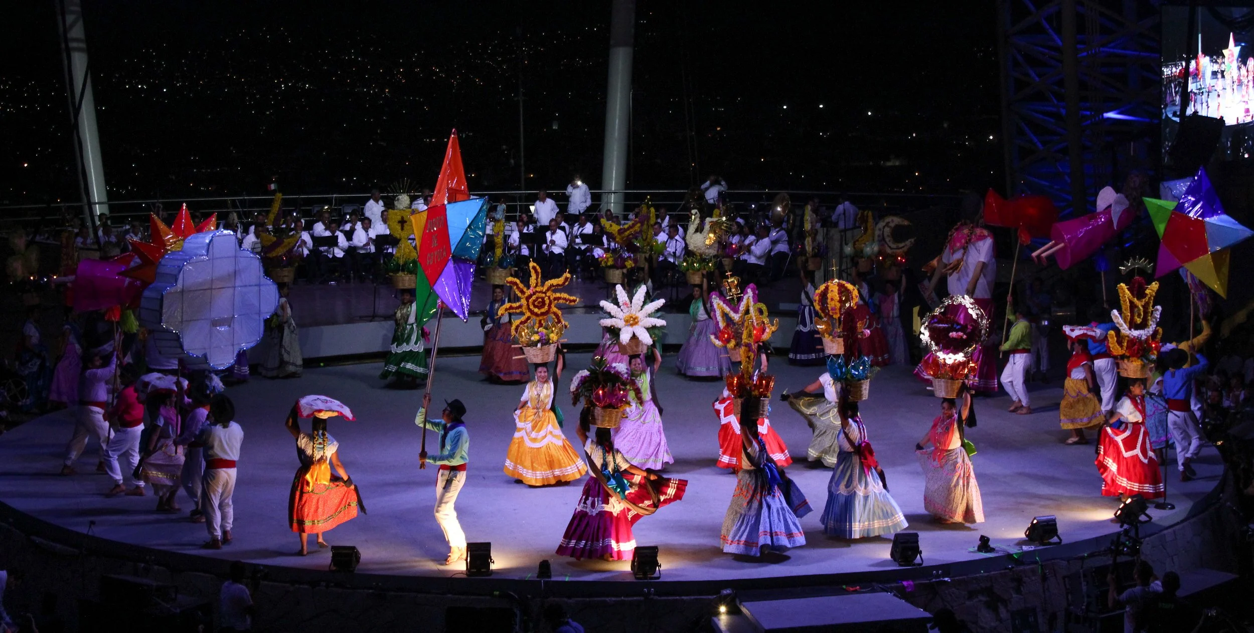 Personas bailando en un escenario durante un espectáculo tradicional con trajes coloridos y un grupo de músicos en el fondo, en una noche con cielo estrellado.