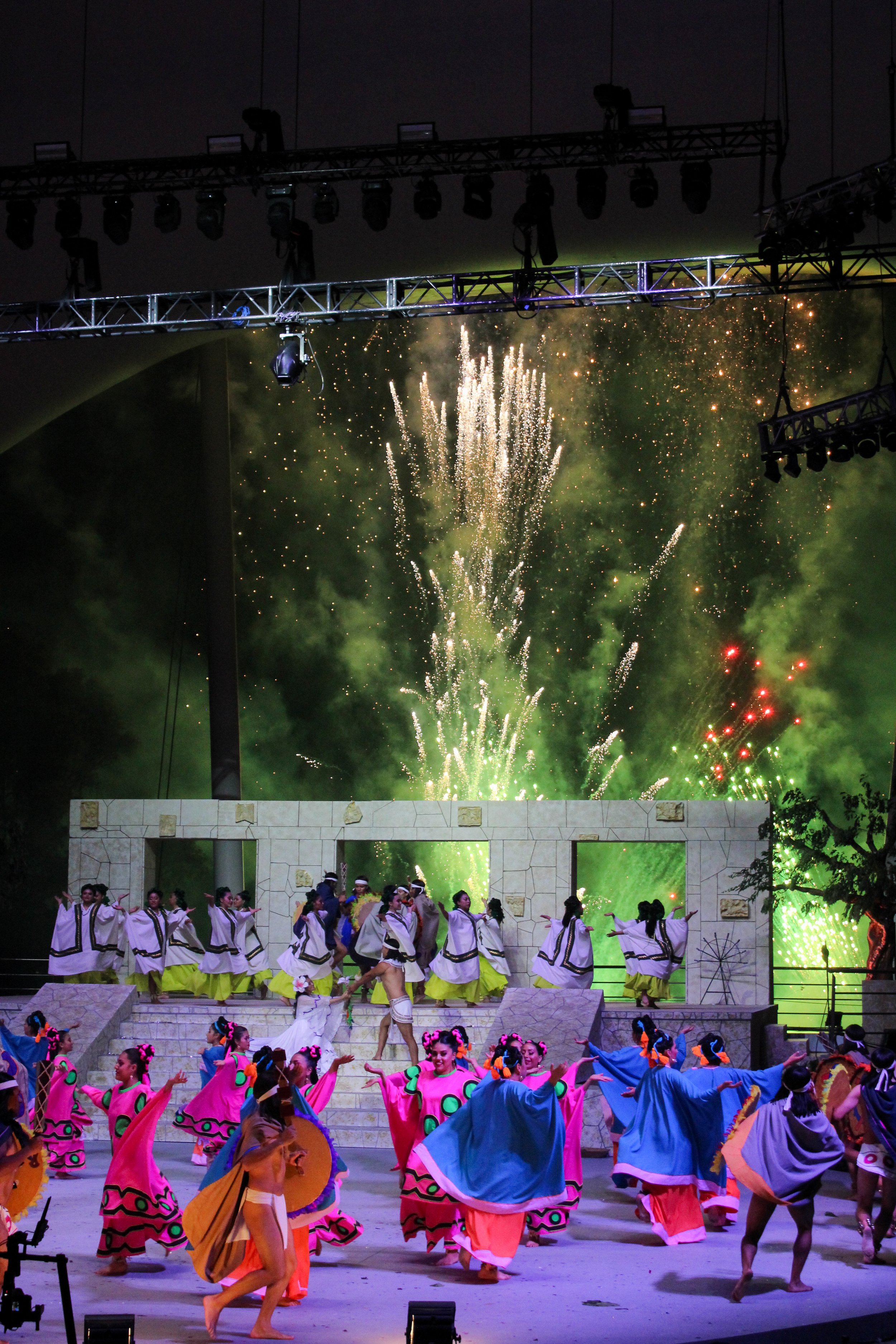 Conjunto de bailarines folklóricos en un escenario con fuegos artificiales en el fondo, participando en una presentación cultural en la noche.