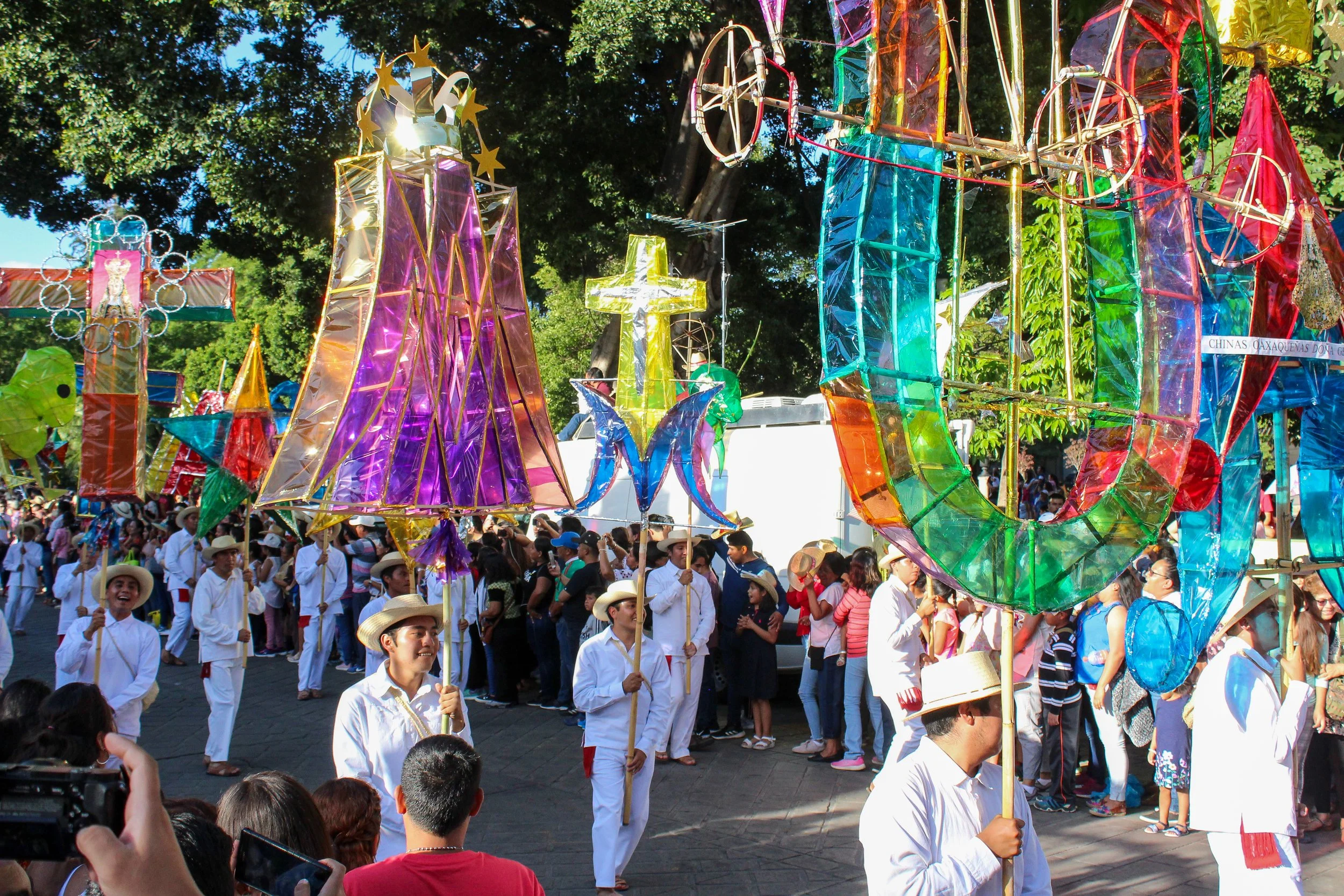 Personas participando en un desfile con faroles rústicos decorativos de colores y formas variadas, muchos llevan sombreros y ropa tradicional en un día soleado con árboles verdes alrededor.