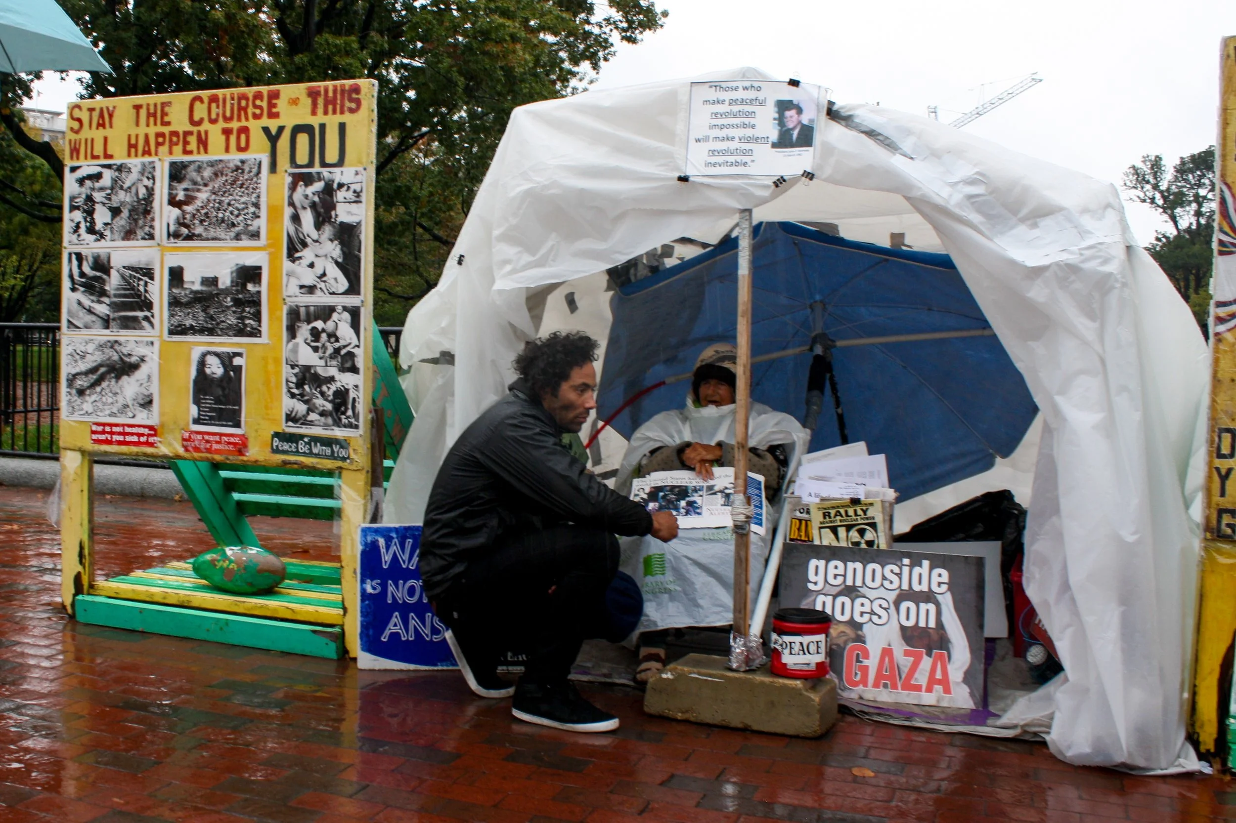 Persona vendiendo en una protesta sobre Palestina, con carteles y productos relacionados con la causa, bajo una estructura temporal en un día lluvioso.