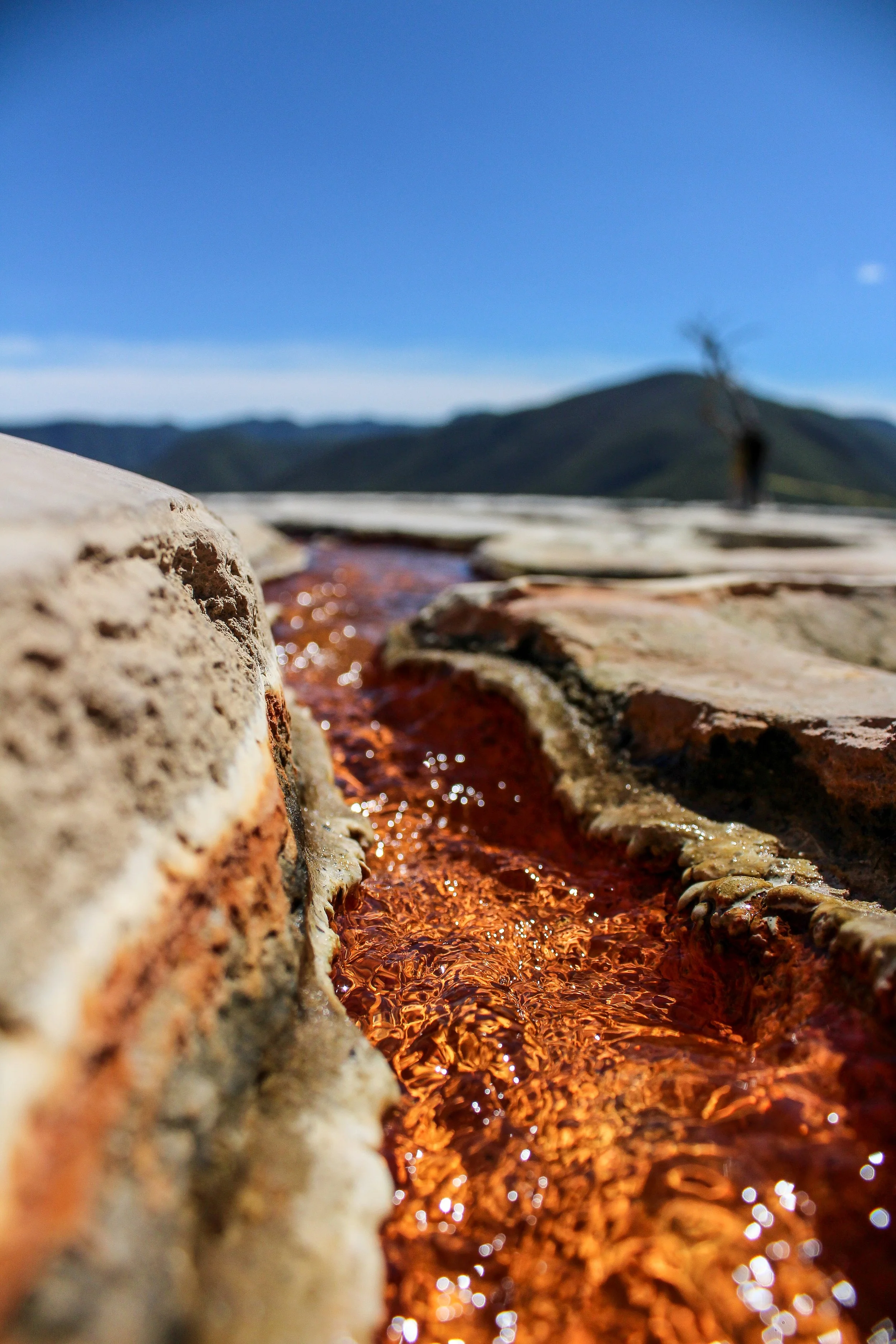 Río de agua caliente o minerales en un paisaje montañoso, con cielo azul y montañas al fondo.