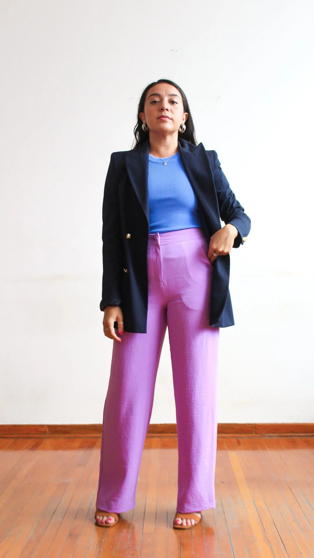 Mujer con pantalones lila, blusa azul y blazer negro, de pie sobre piso de madera, mirando hacia la cámara.