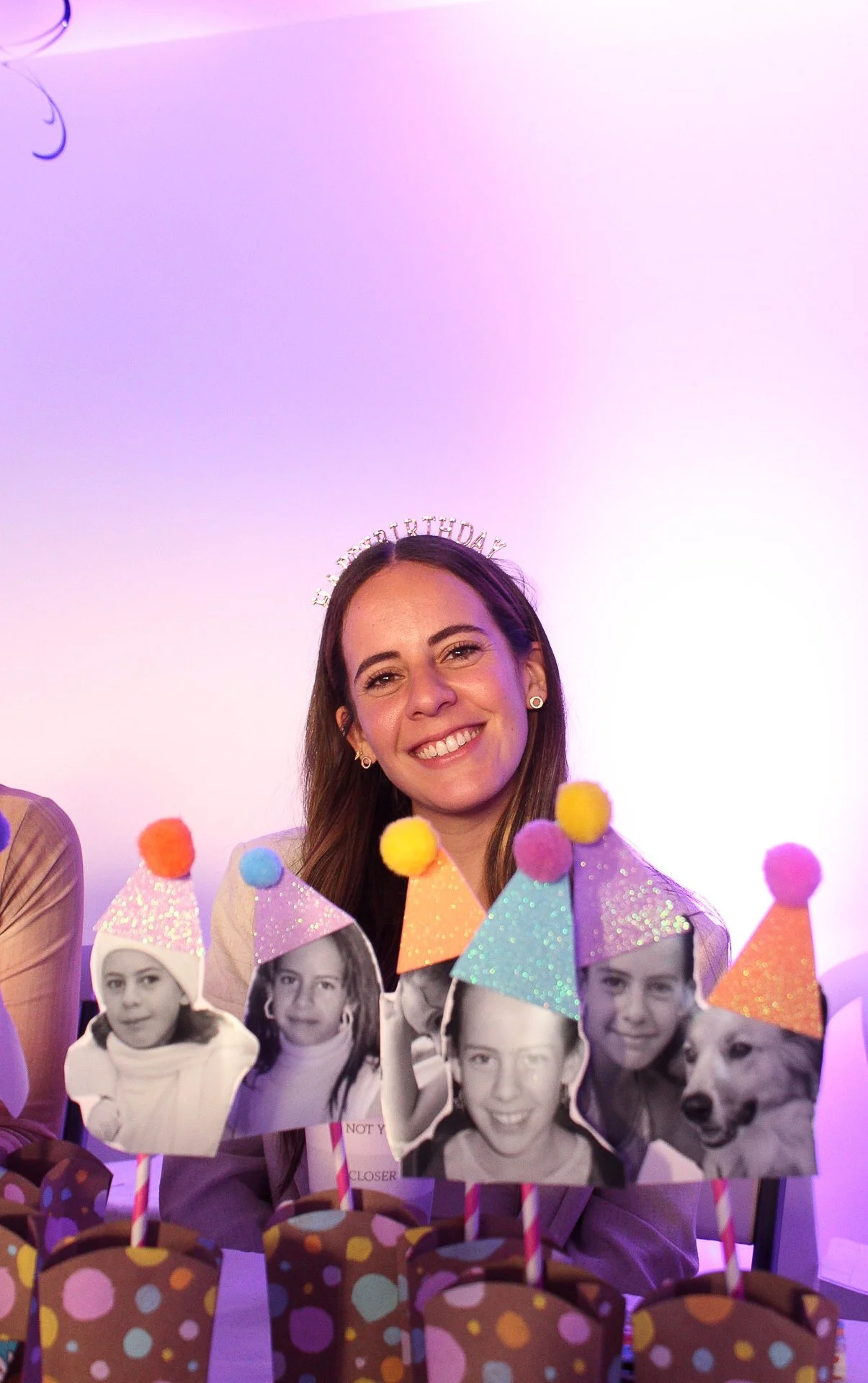 Mujer sonriendo en fiesta de cumpleaños con sombreros de fiesta, foto en blanco y negro de niños, perros y mujer, y decoraciones coloridas.