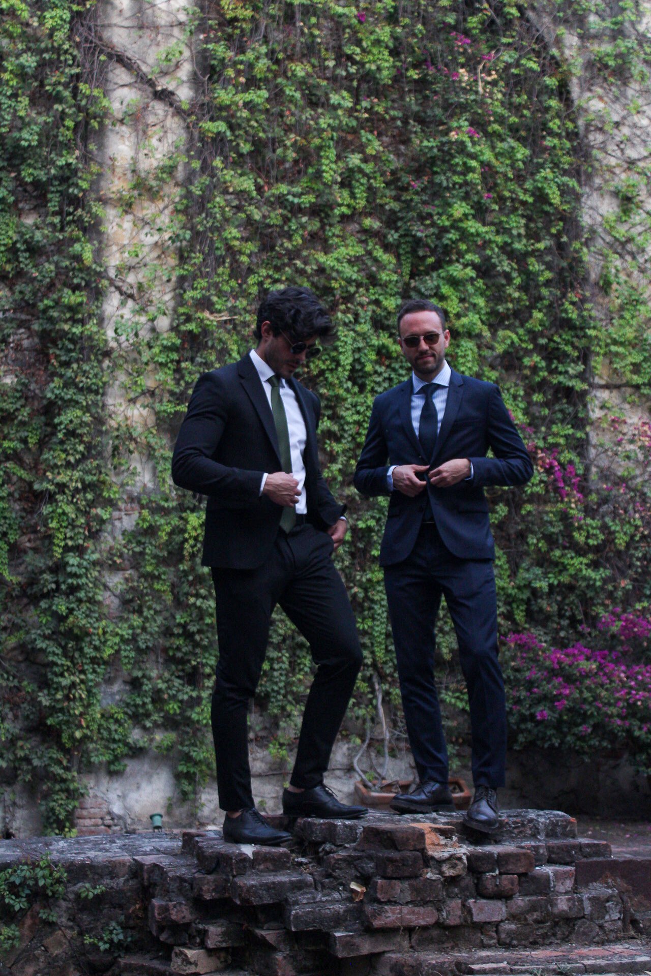 Dos hombres con trajes oscuros y gafas de sol, de pie en una estructura de ladrillos, en un fondo de enredaderas verdes y flores moradas.