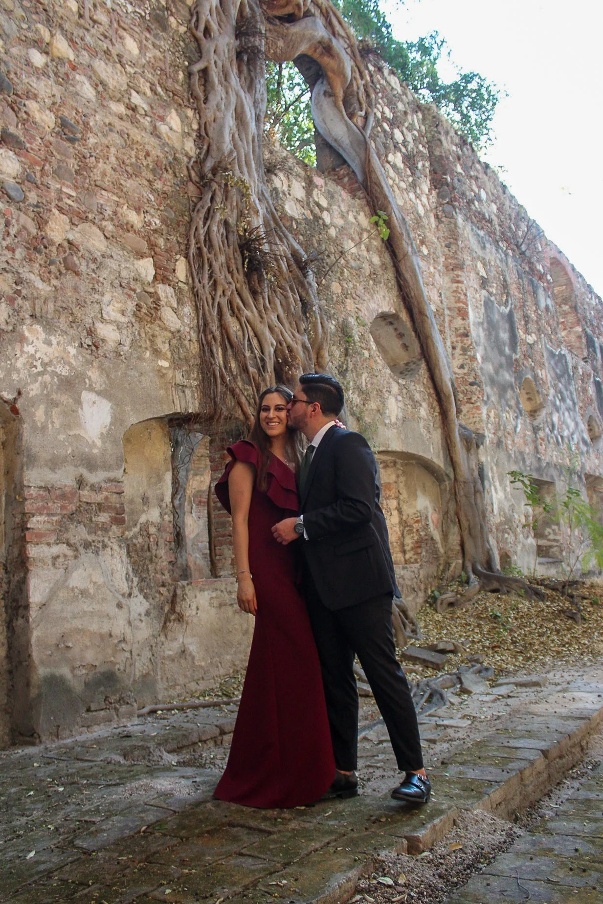 Una pareja vestida formalmente, la mujer con vestido largo burdeos y el hombre con traje negro, frente a una pared antigua de ladrillos con raíces de árbol colonizando la estructura, en un entorno exterior.