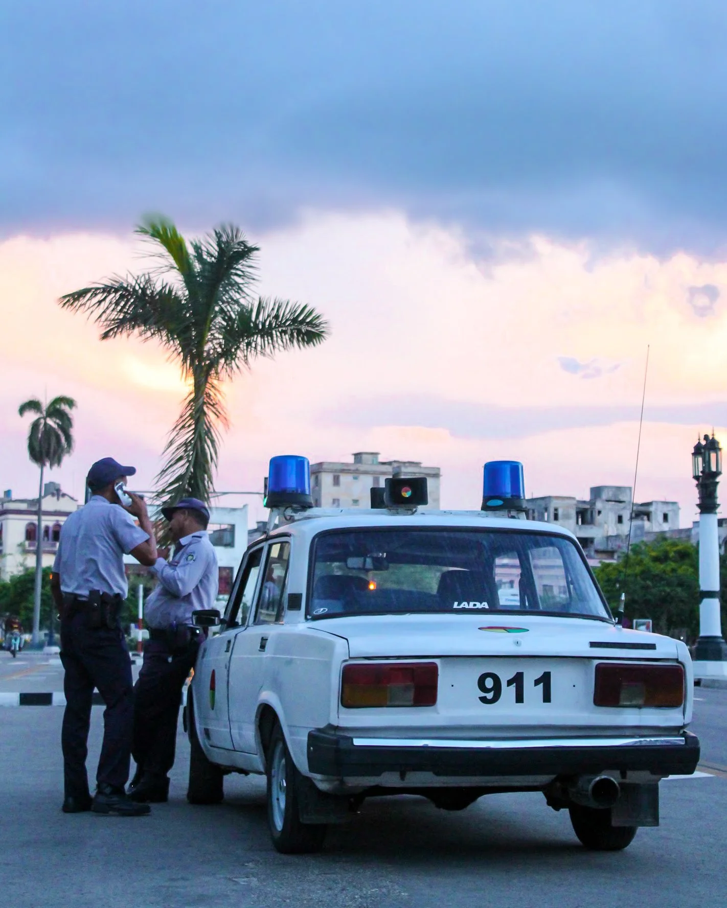 Dos oficiales de policía conversan junto a un coche patrulla en una plaza con palmeras y edificios en el fondo, durante el atardecer.