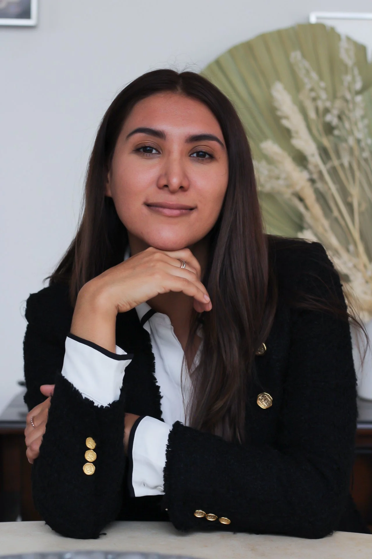 Retrato de una mujer joven con cabello largo y oscuro, vestida con ropa formal negra y blanca, sonriendo suavemente, apoyando la barbilla con la mano, frente a un fondo decorativo con una planta grande y hojas secas.